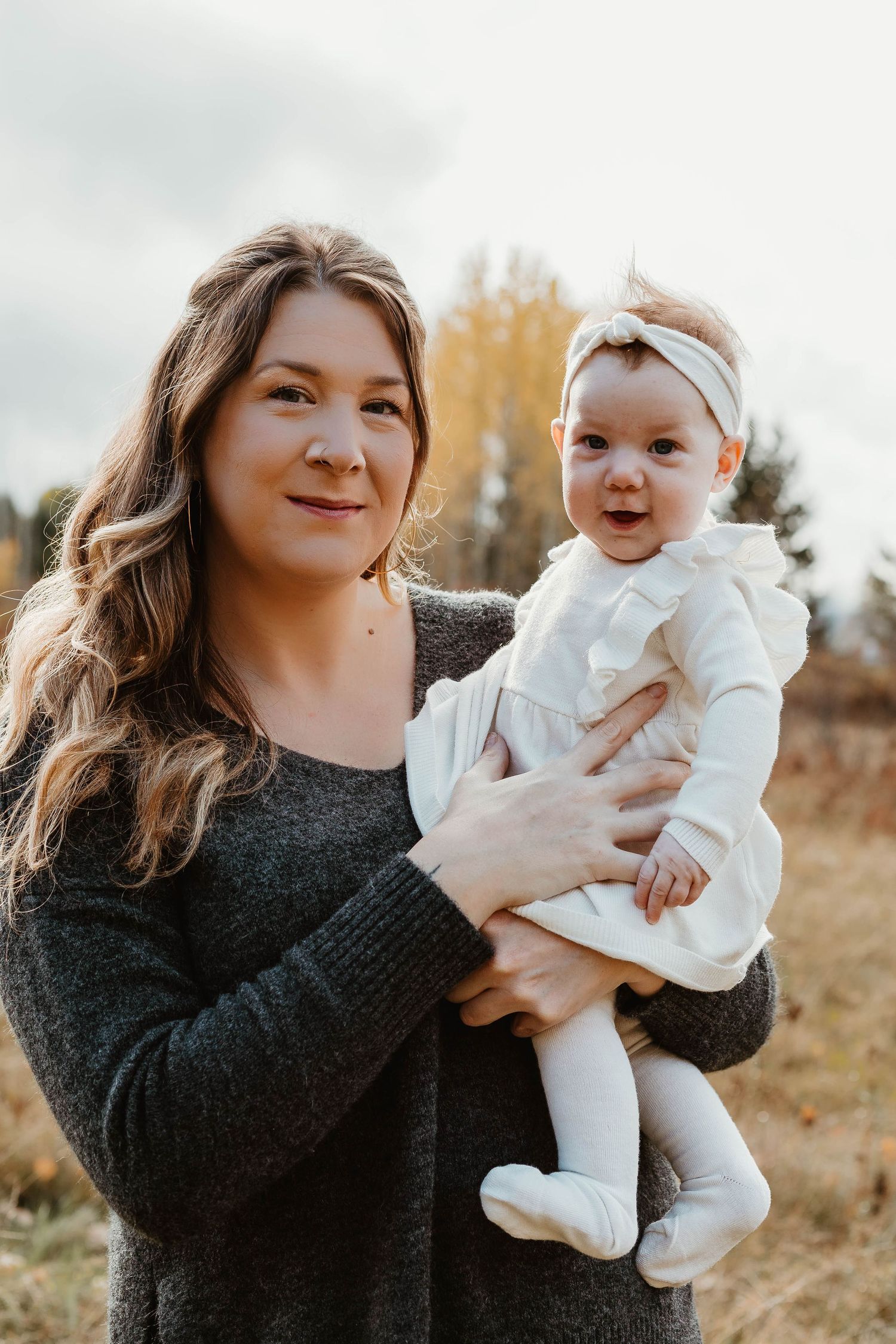 Woman in gray sweater holding happy baby in white dress outdoors during autumn.