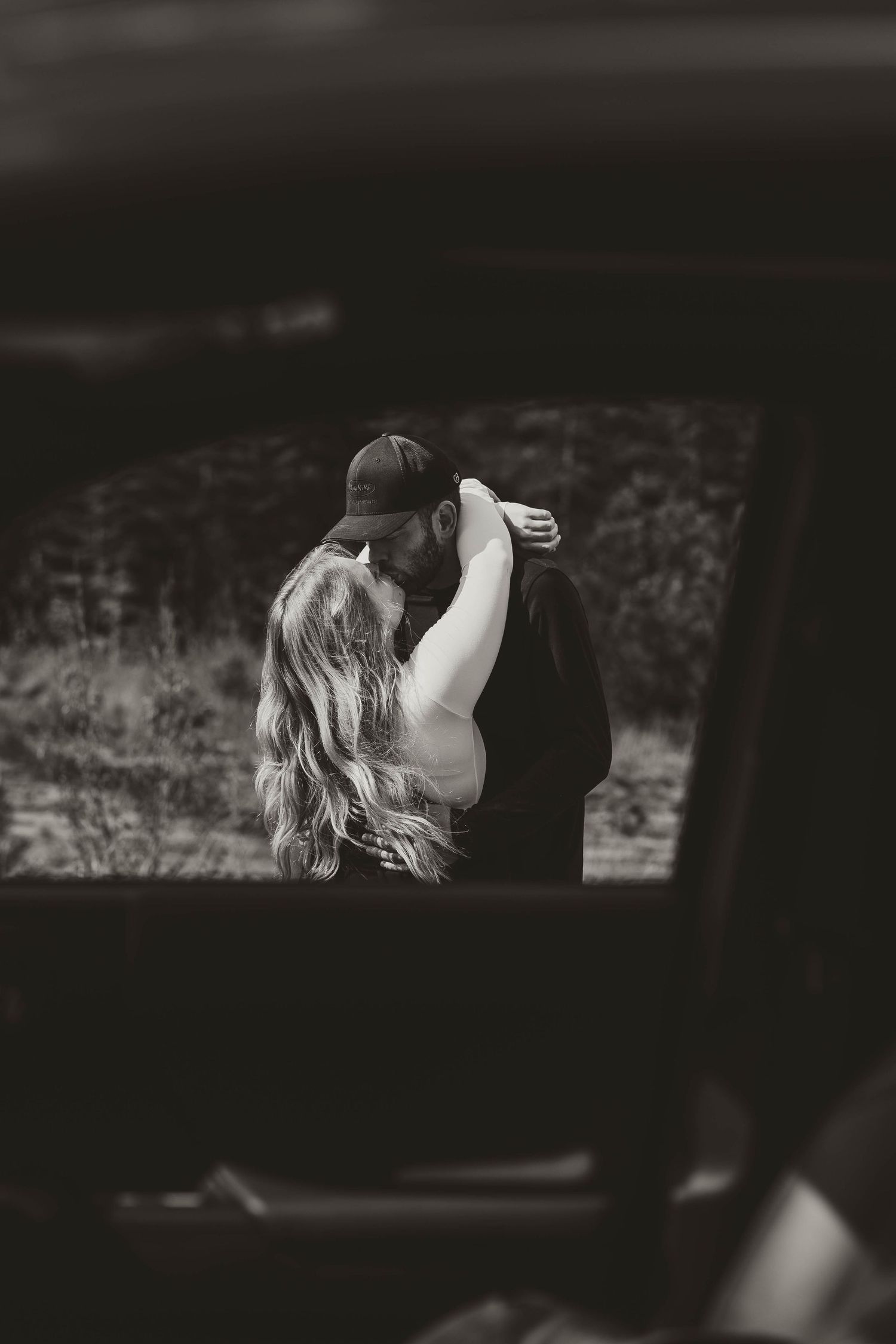 A romantic black and white photograph of a couple embracing through a car window frame.