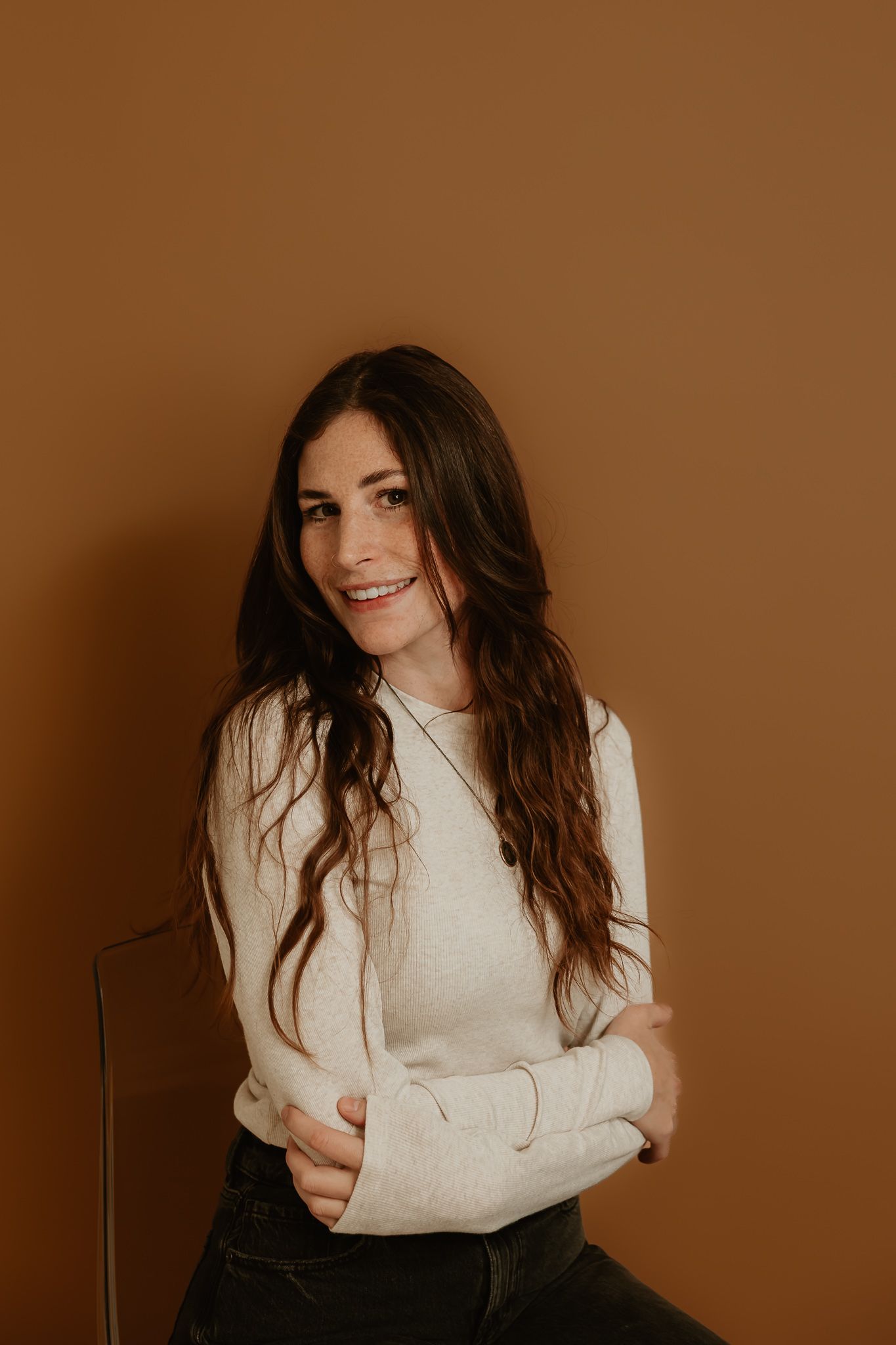 A person in a cream sweater poses against a brown background with long dark hair and a playful smile.
