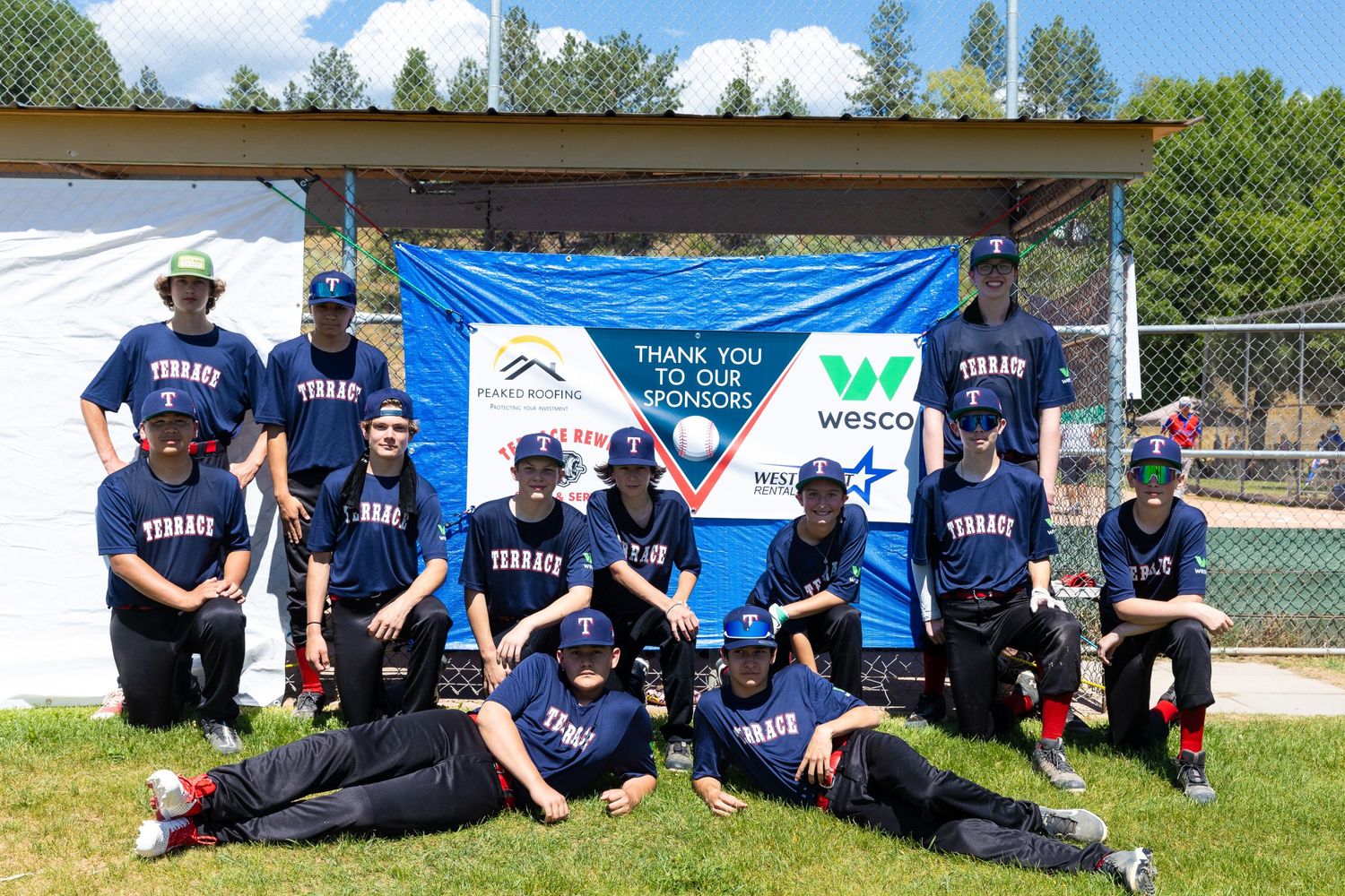 Youth baseball team in navy blue uniforms poses for a group photo under a banner at their field.