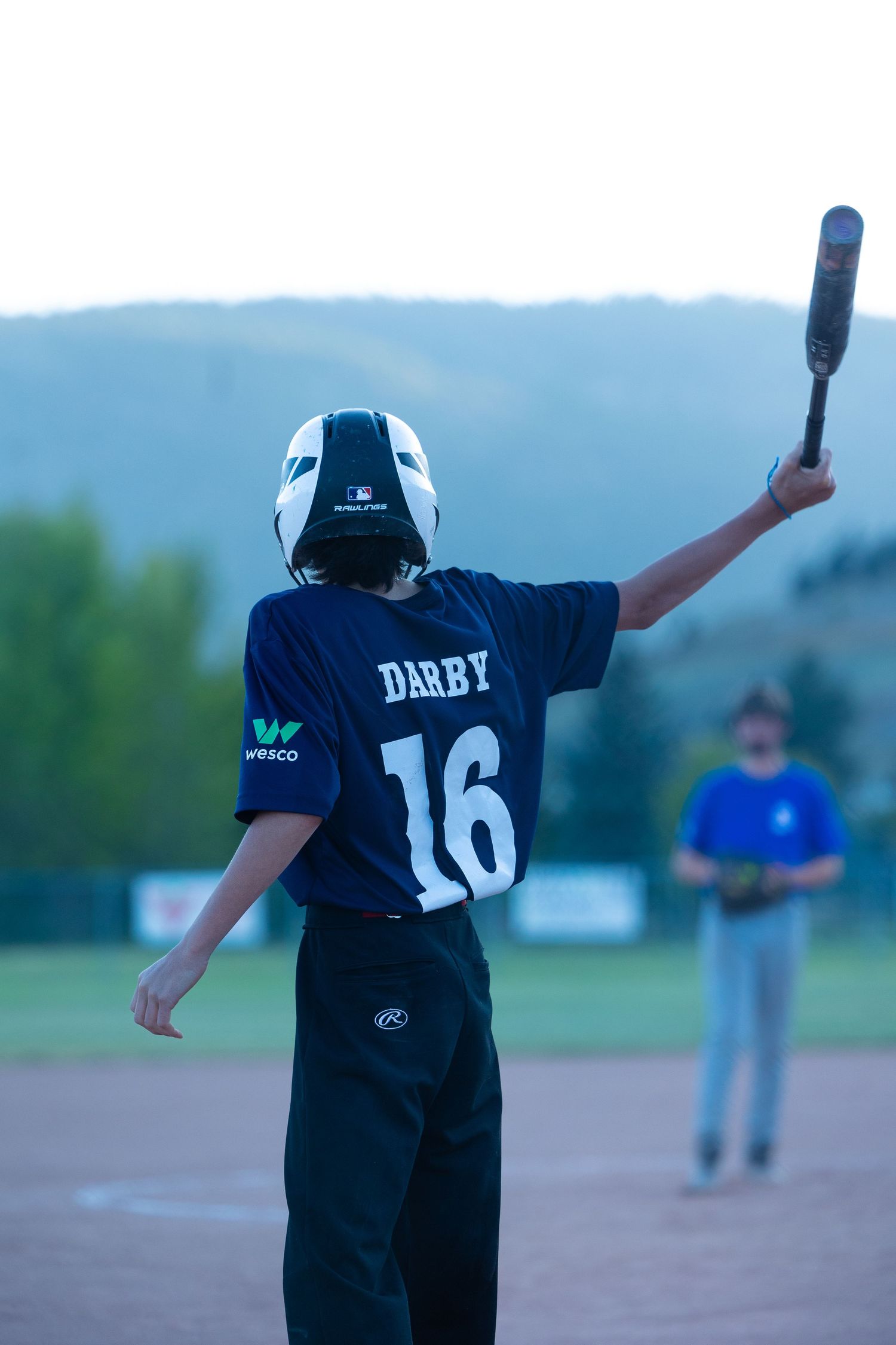 Baseball player wearing navy jersey number 16 holds up a bat on the field at dusk.