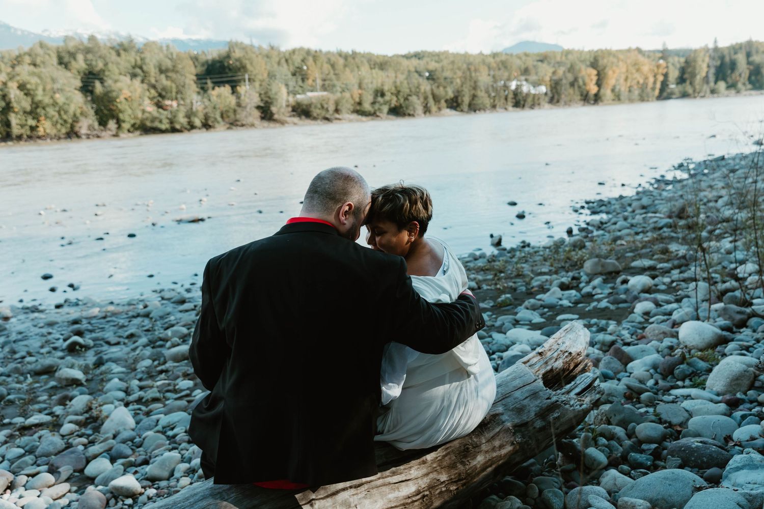 Couple sits together on driftwood log by rocky riverbank during sunset.