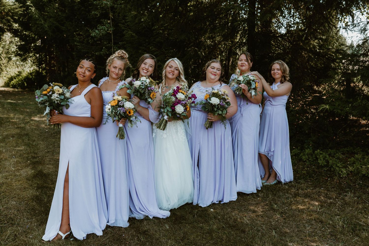 Bridesmaids in flowing lavender dresses pose with bouquets against forest backdrop.