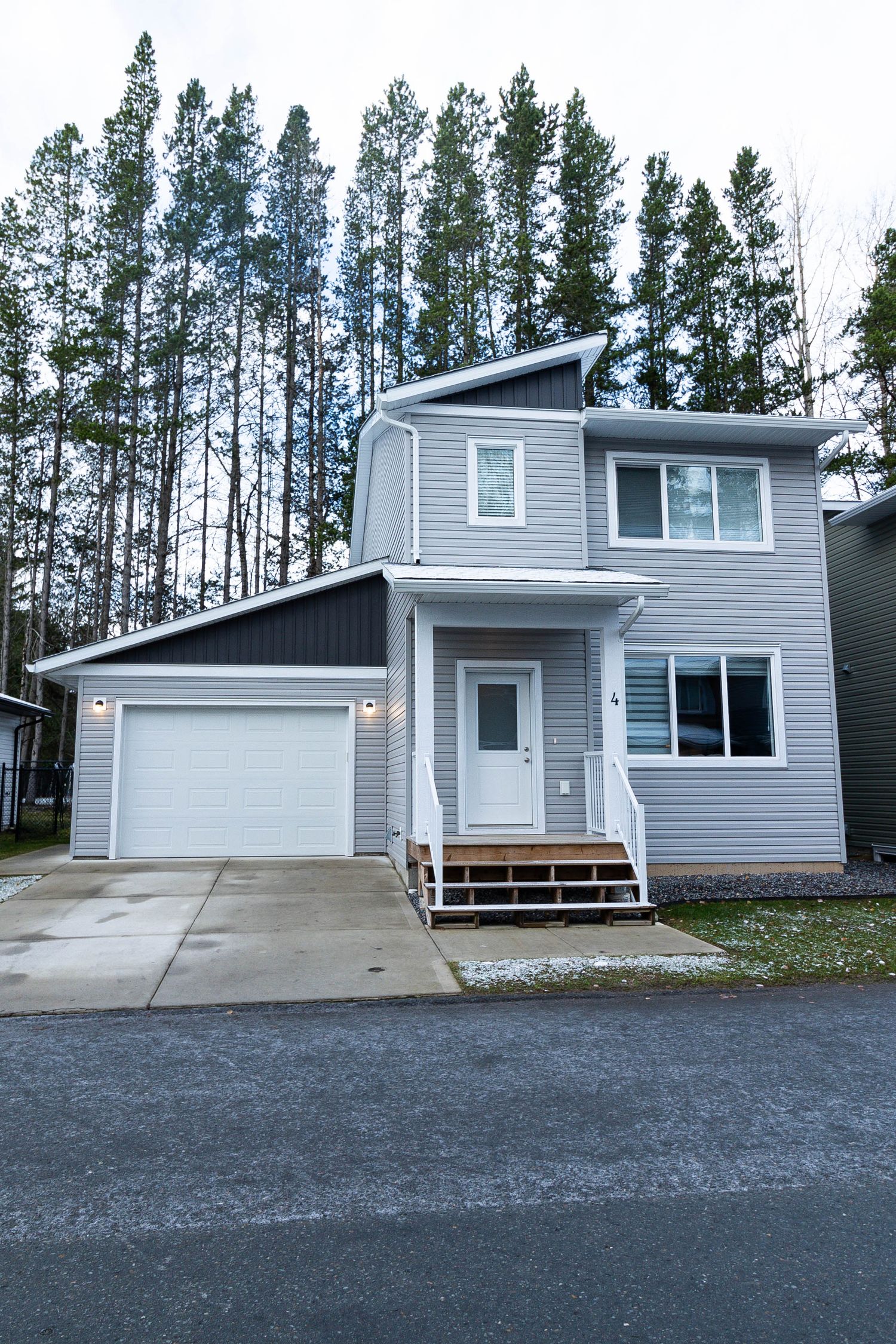Modern gray two-story house with attached garage and tall pine trees in background.