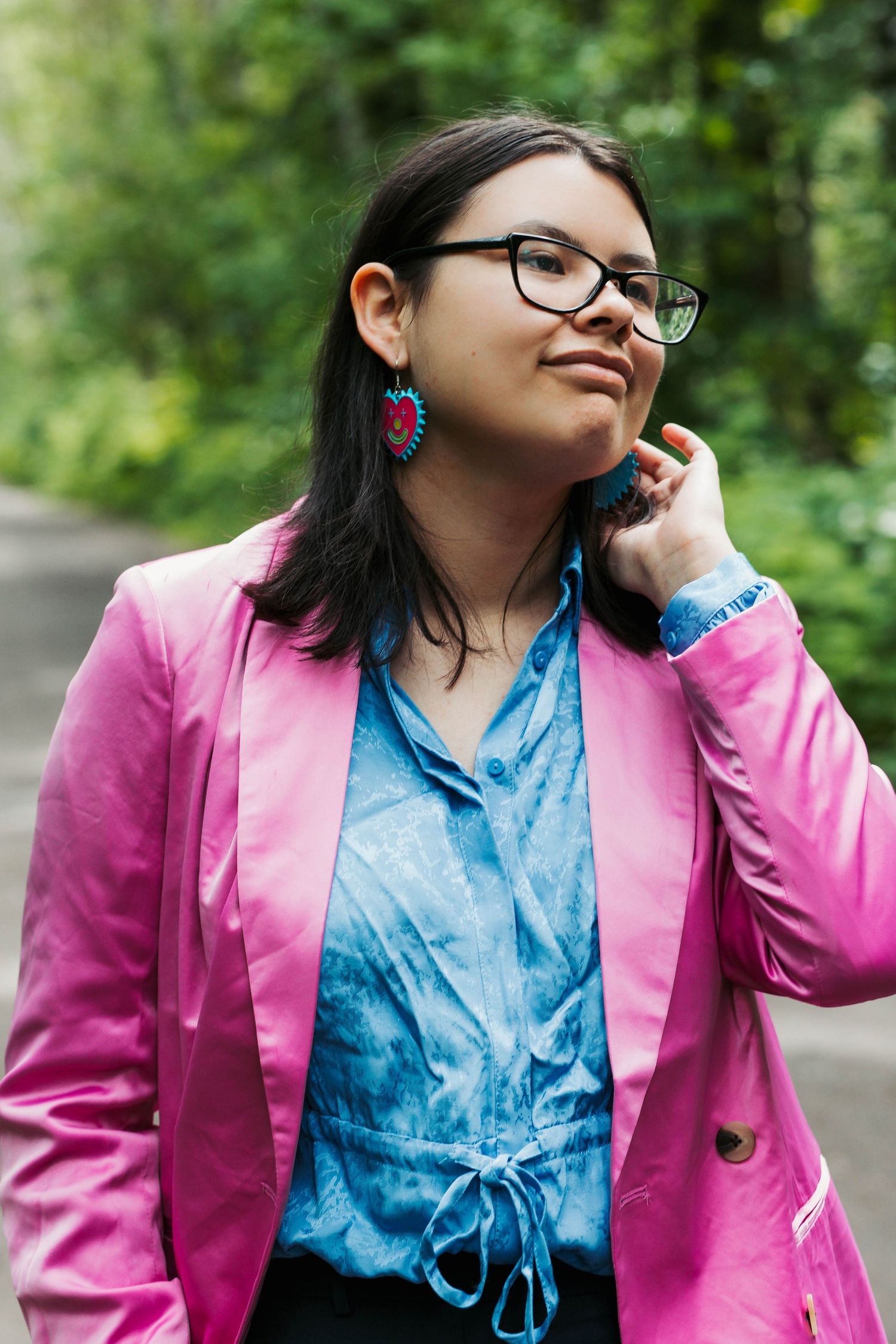 Professional portrait of person wearing a pink blazer and blue top with colorful earrings against a green outdoor background.