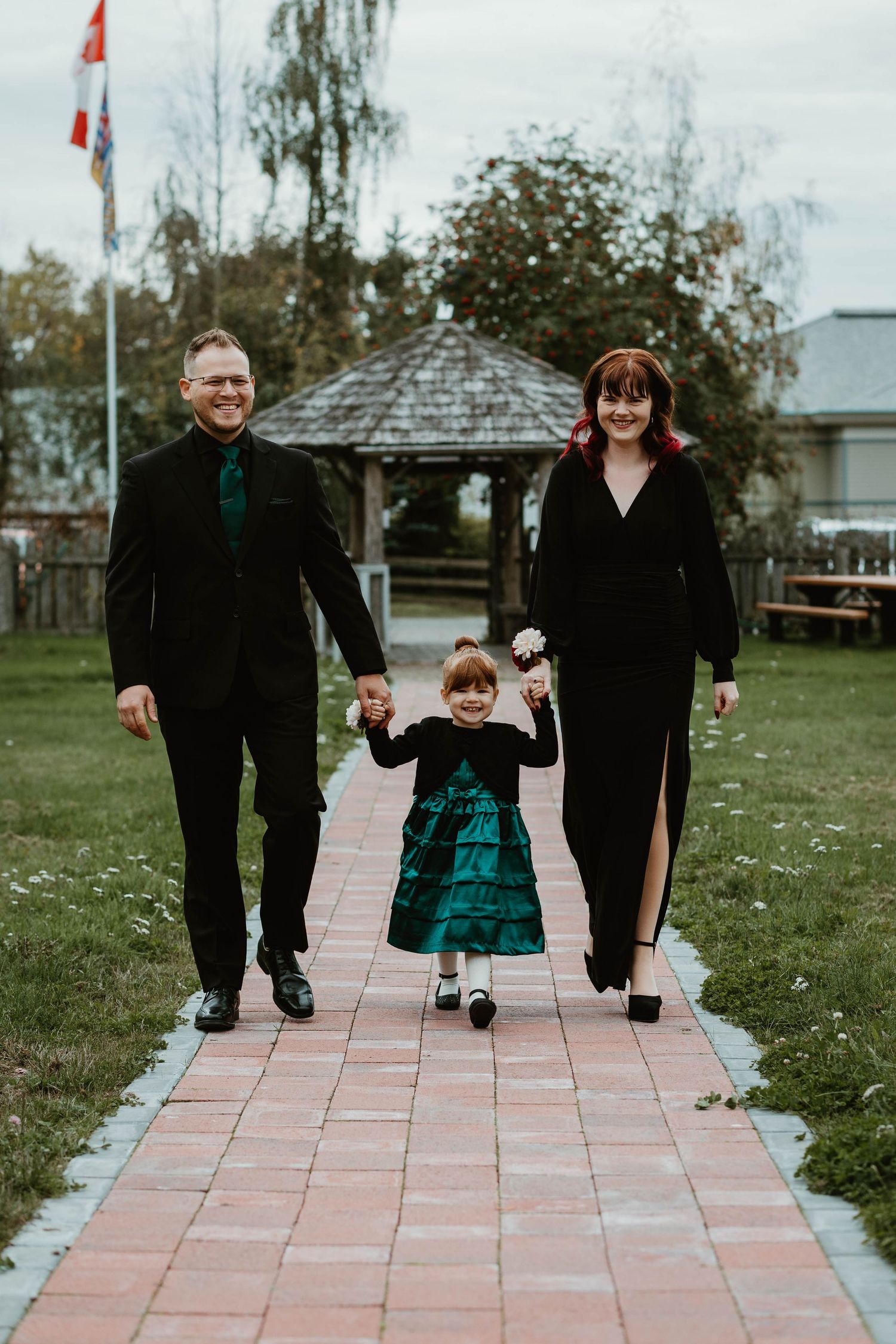 A child in a teal dress walks down a brick path holding hands with two adults wearing black formal attire.