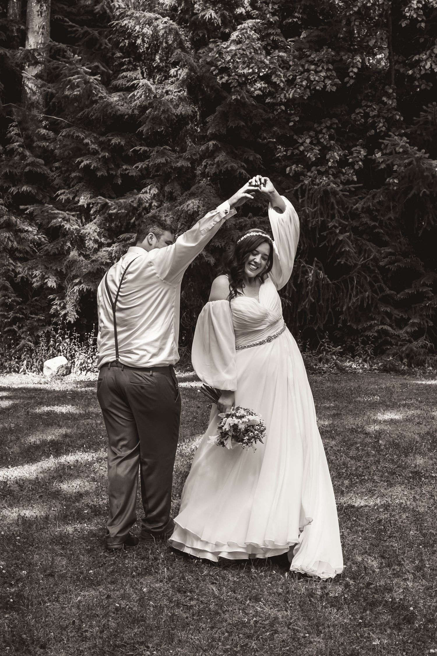 Black and white photo shows couple dancing playfully in a forest clearing on their wedding day.