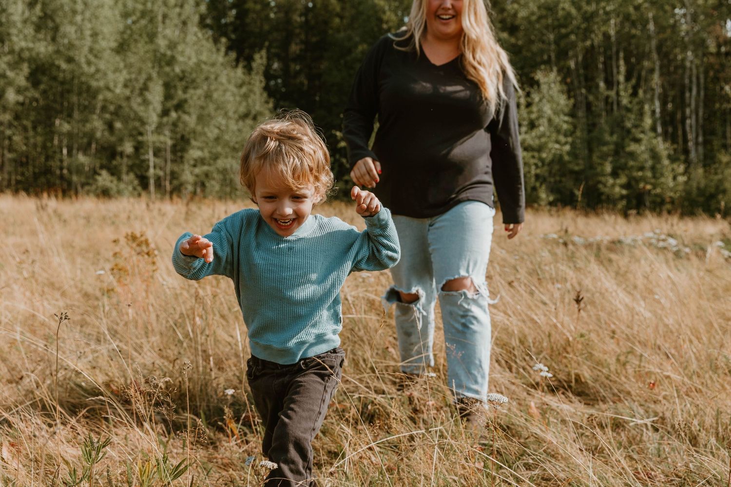 A toddler in a blue sweater runs through a golden field while holding hands with someone wearing jeans.