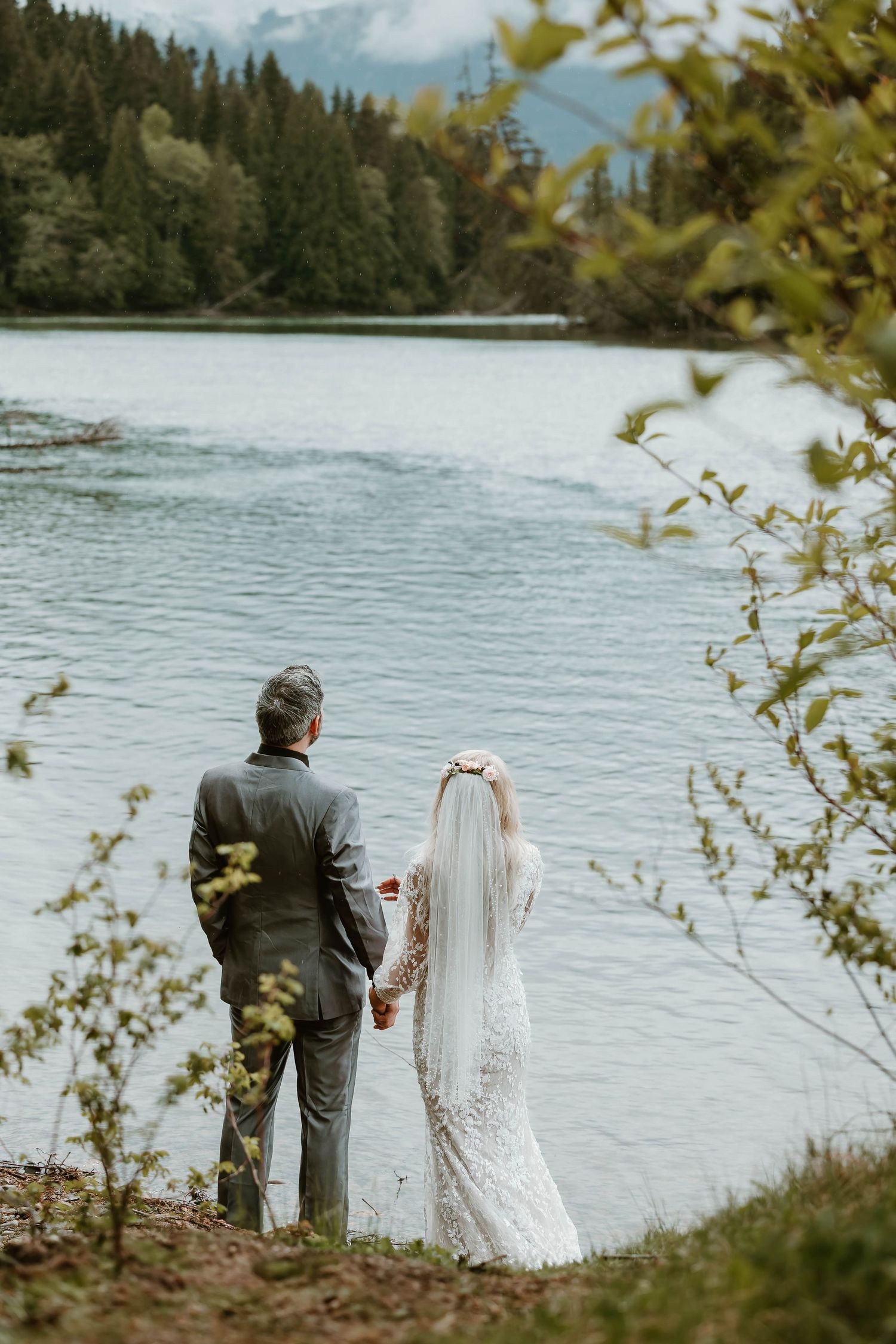 A couple in formal attire stands by a serene mountain lake surrounded by evergreen trees and cloudy skies.