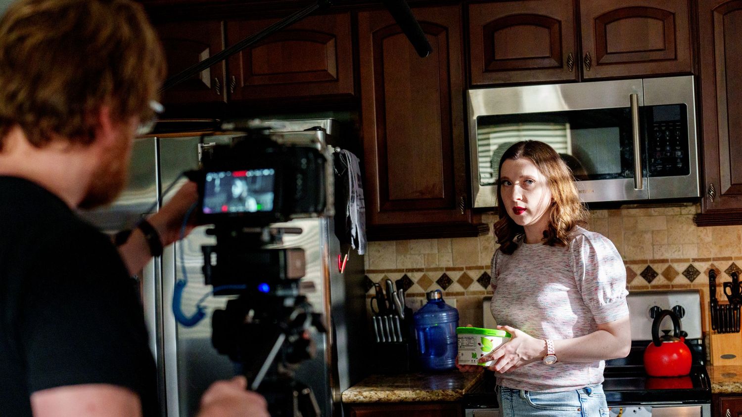 Behind-the-scenes filming in a kitchen shows a camera operator recording someone holding food items near wooden cabinets.