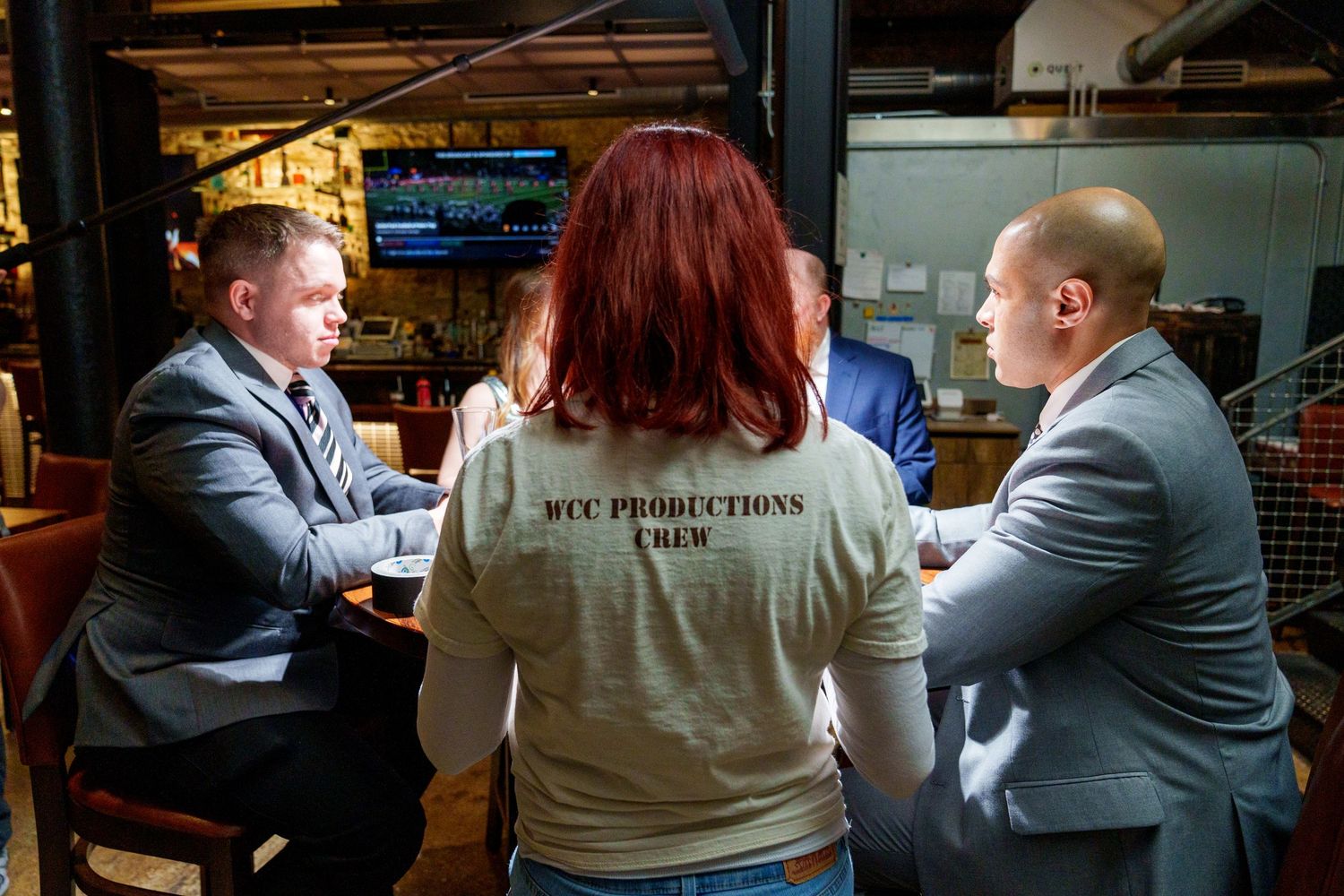 MCC Productions crew member in branded shirt meets with two men in suits at a restaurant table.