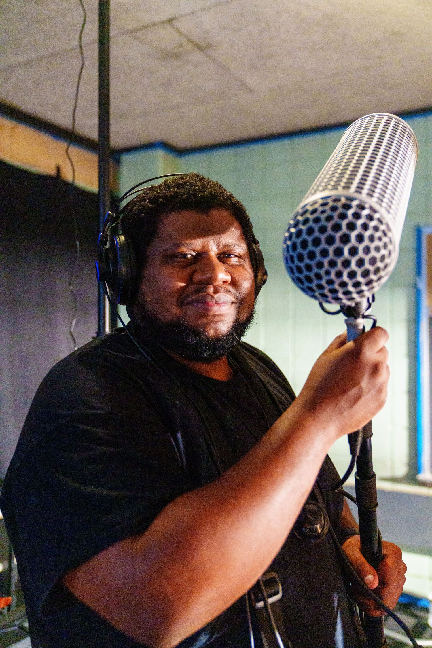Smiling man with headphones holds a vintage studio microphone in a recording studio.