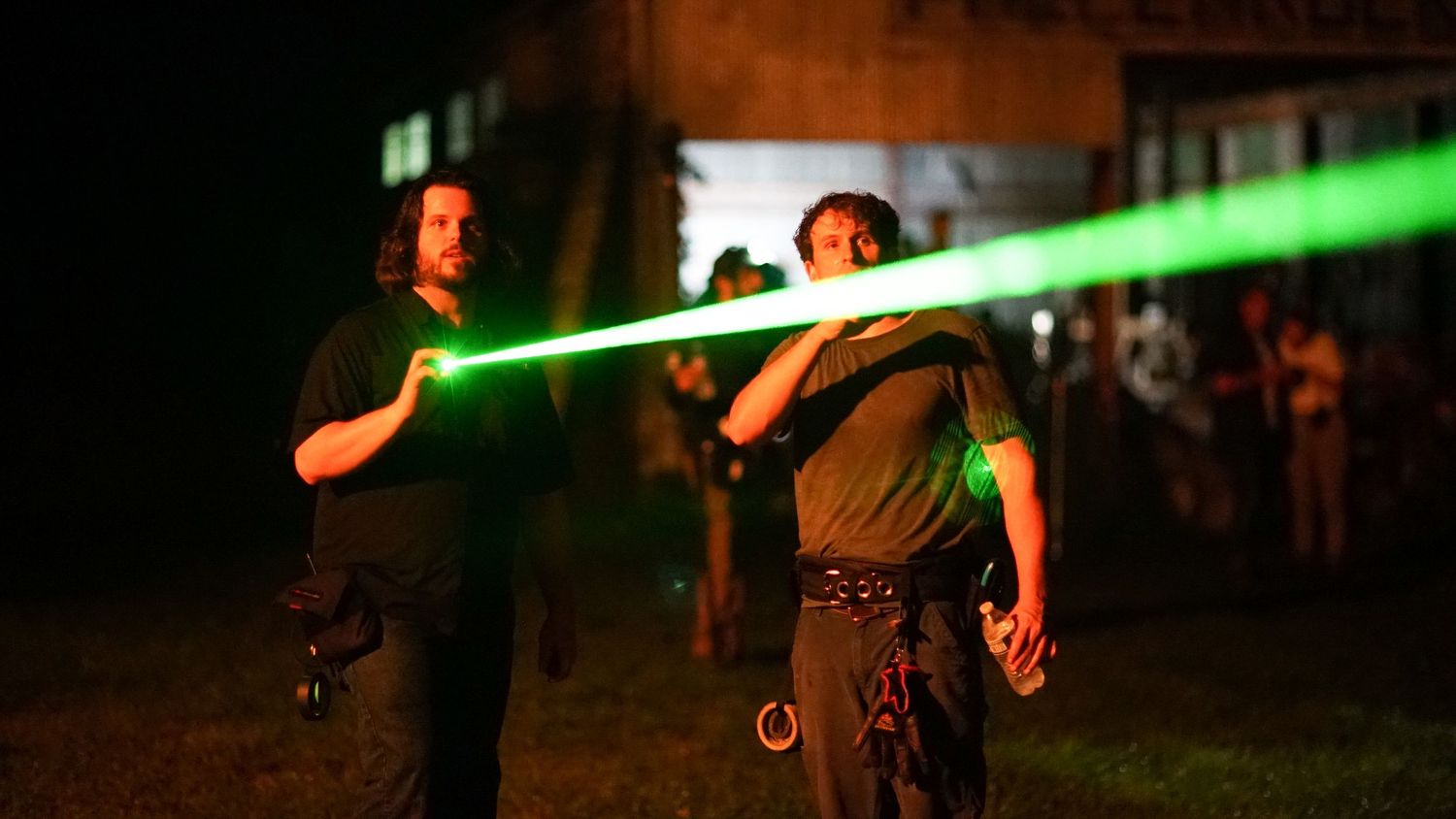 Two people holding bright green laser beams at night outdoor event with crowd in background.