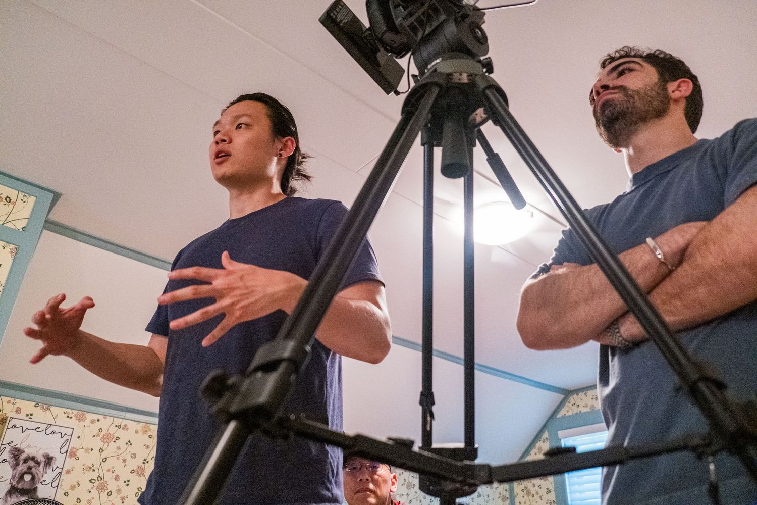 Filmmaker gesturing near a camera on tripod while collaborating with crew member indoors.