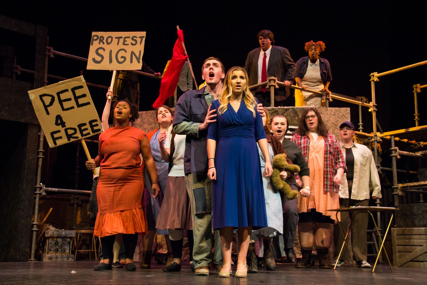 Theater performers hold protest signs during stage production scene.