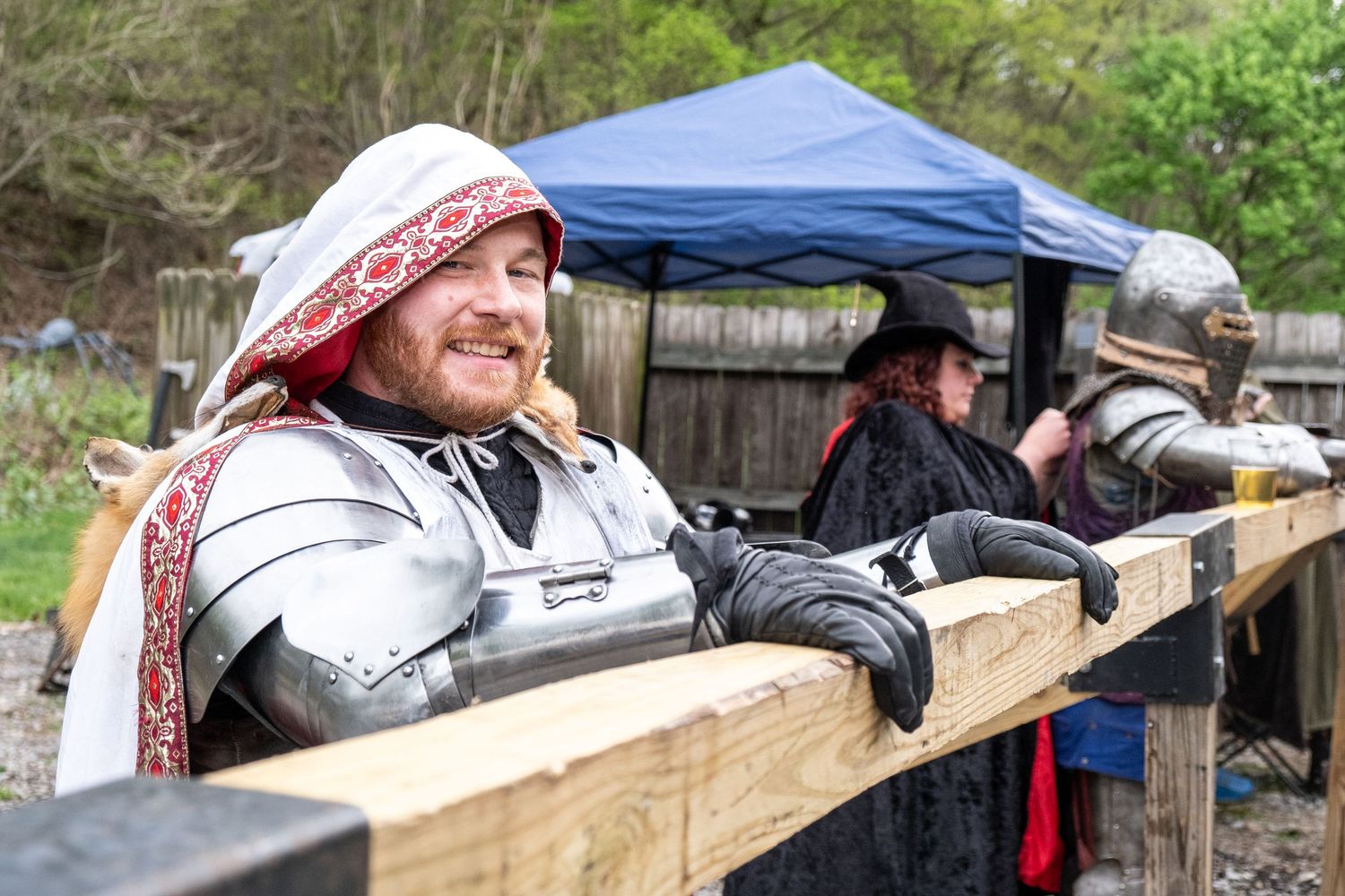 People in medieval costumes relaxing at wooden fence during outdoor event.