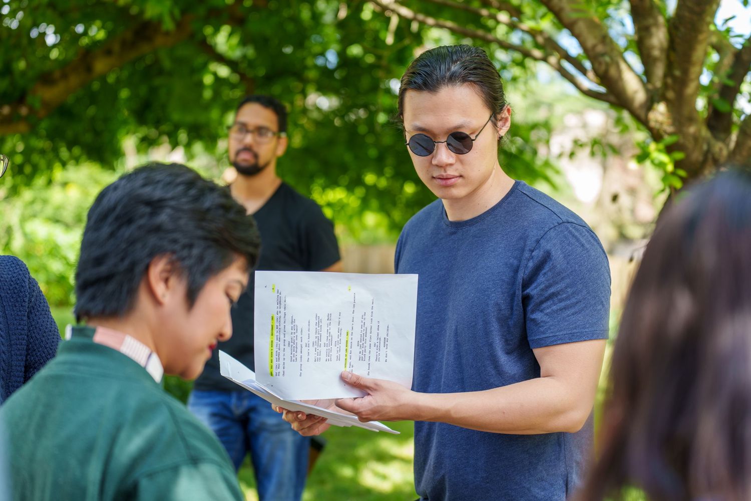 A group of people gather outdoors under trees having a casual conversation.