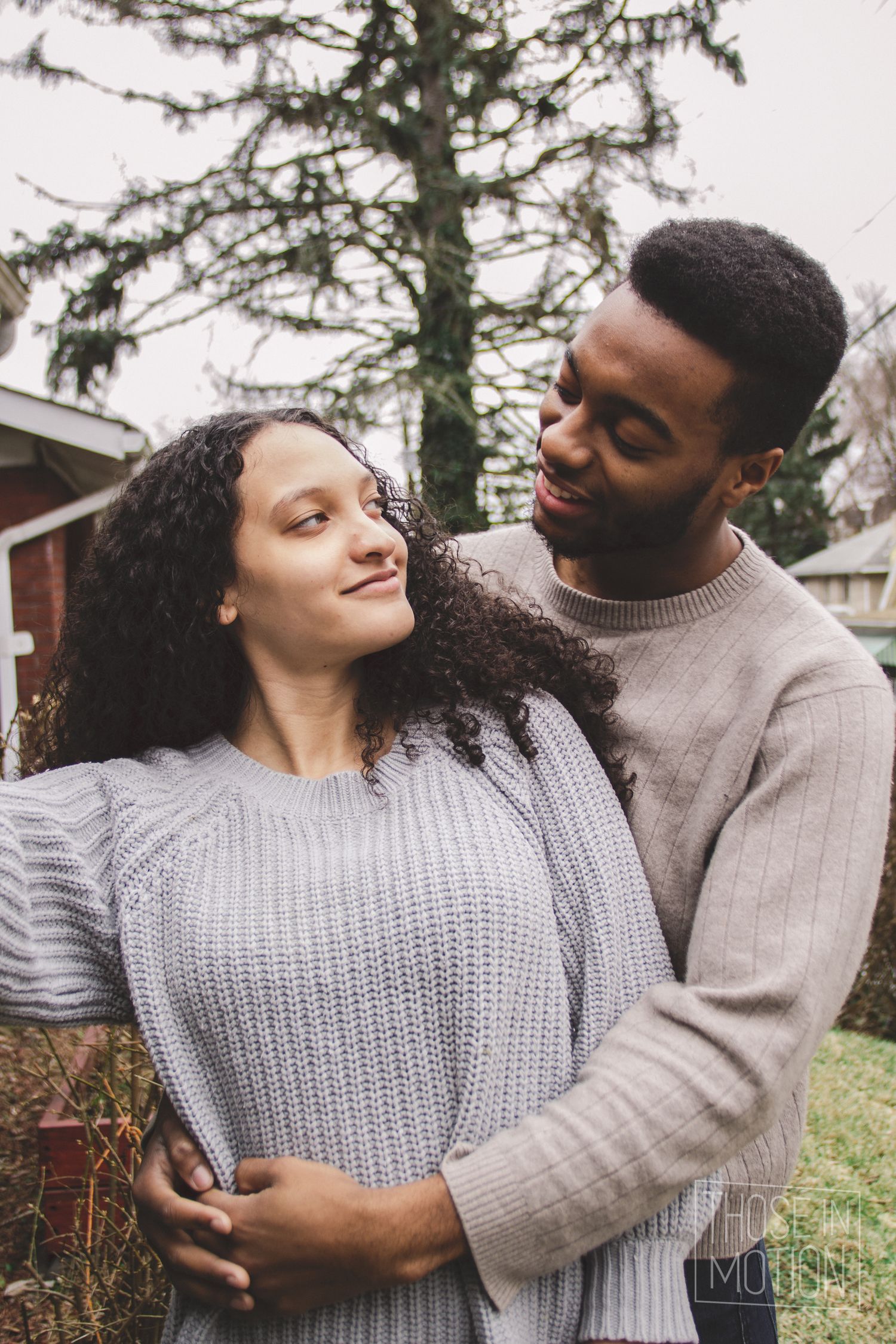 Couple in knit sweaters embracing outdoors against evergreen trees.