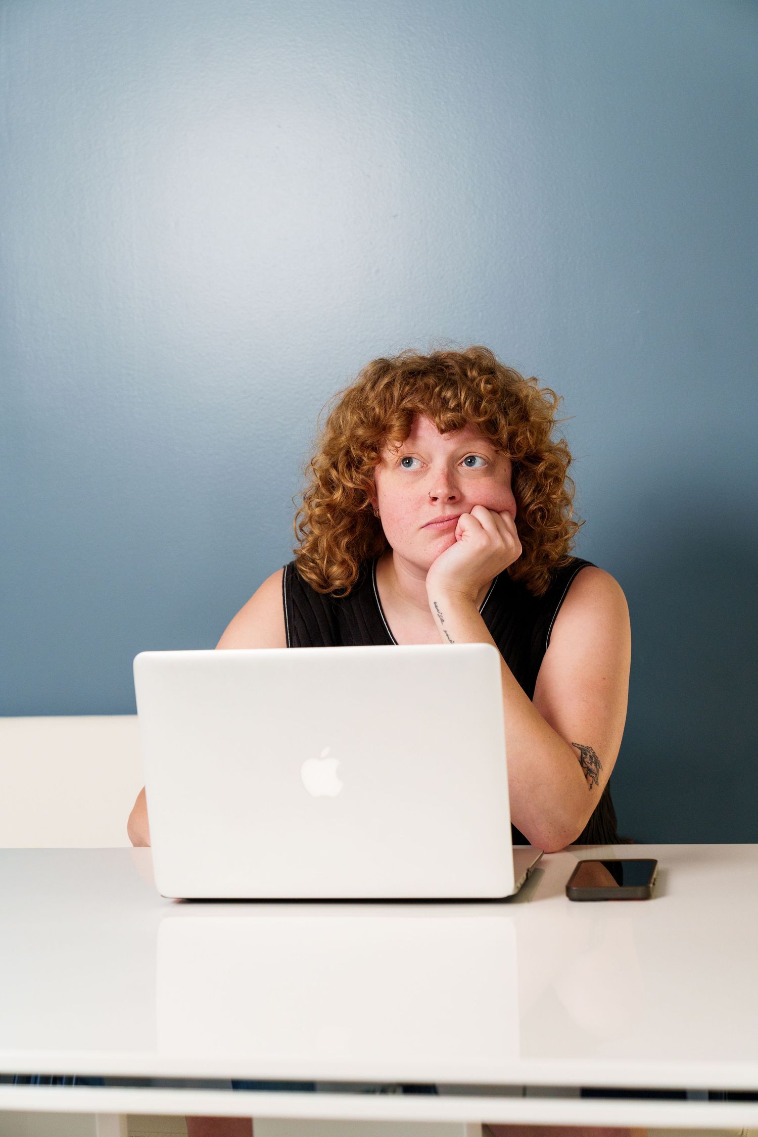 Person with curly hair looking bored while working on MacBook laptop at white desk.