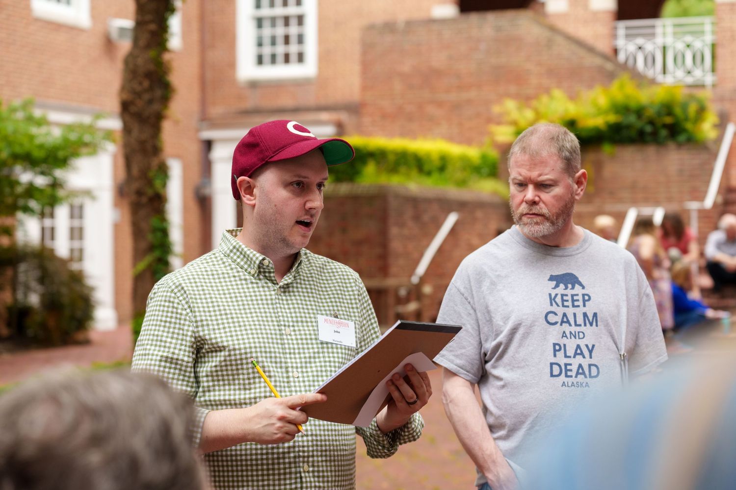 Two men outdoors, one holding clipboard wearing red cap, other in Keep Calm Play Dead shirt, at casual event.