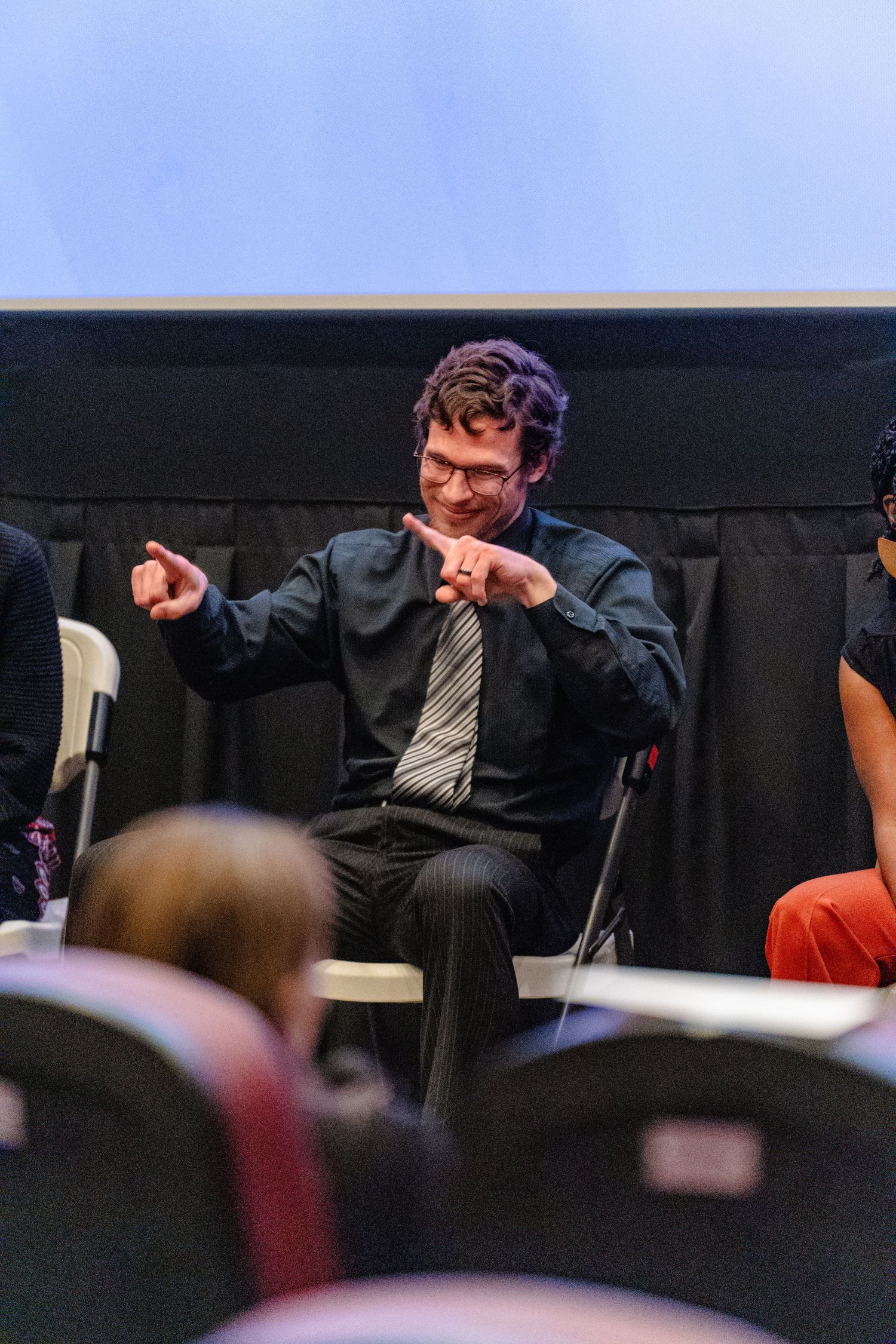 A speaker gestures expressively while presenting to an audience in a darkened auditorium setting.