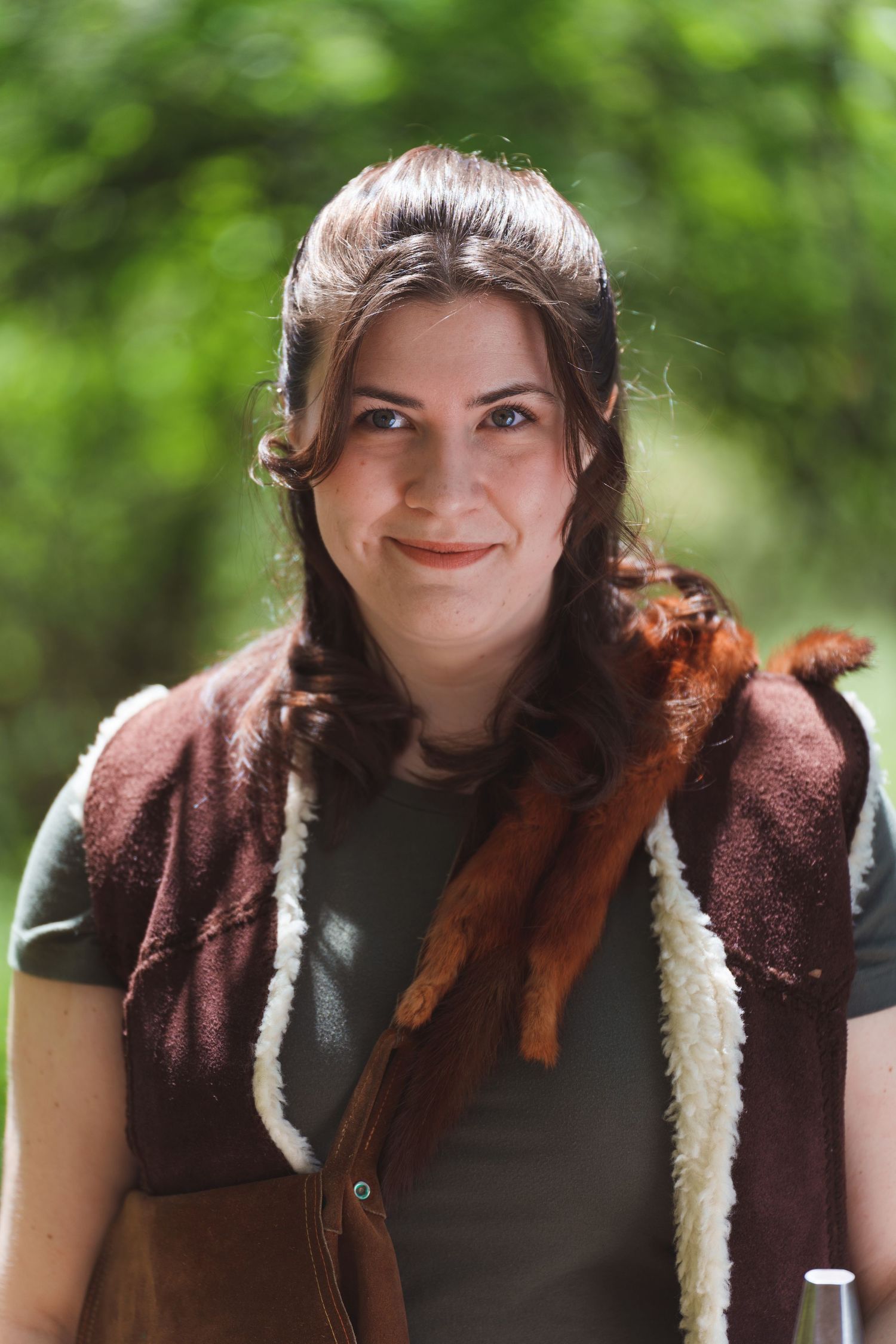 Person wearing a brown vest with fur trim smiles in an outdoor setting.