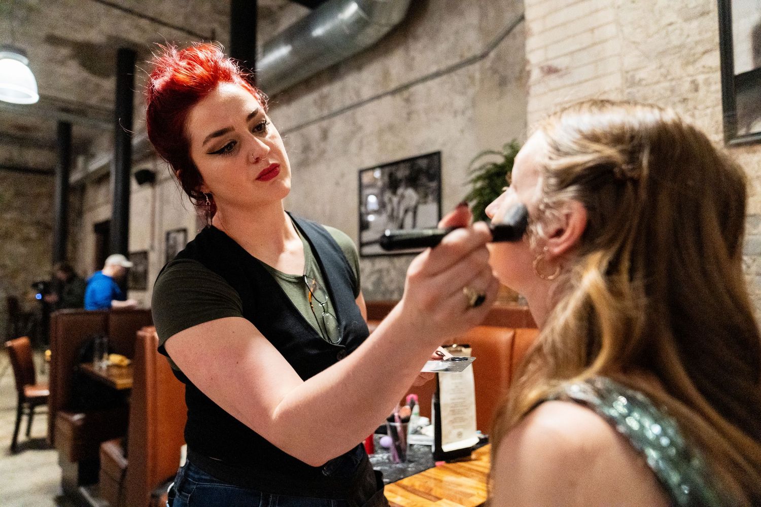 A makeup artist with red hair applies cosmetics to a woman in a rustic brick interior setting.