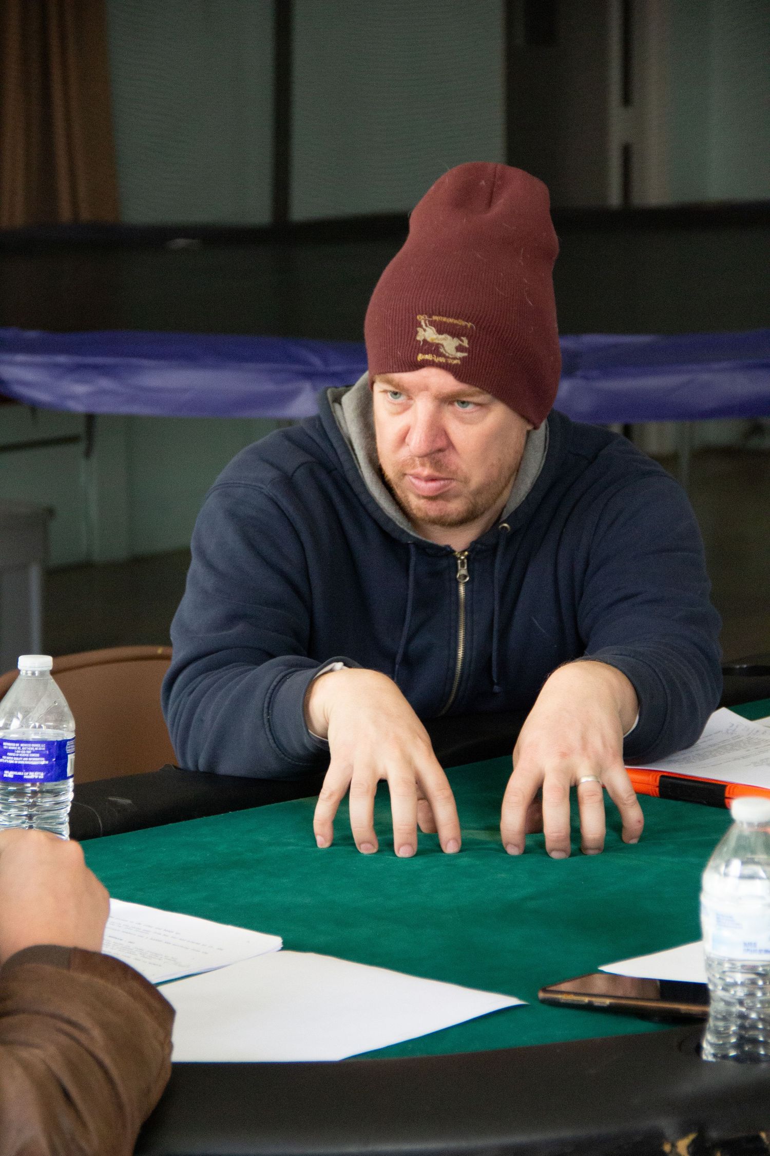 Man in navy hoodie and maroon beanie sits at green felt poker table with hands flat, water bottles nearby.