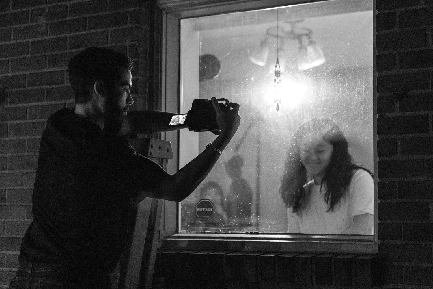 Man photographing woman through foggy window at night in black and white urban setting.