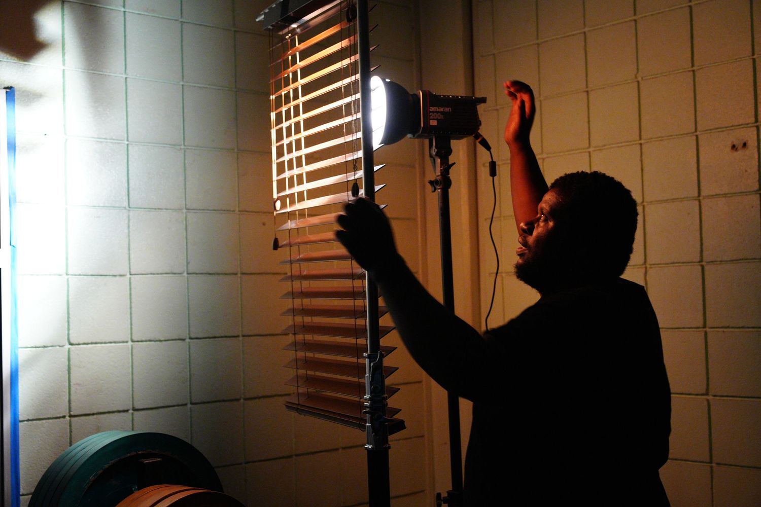A person adjusts a bright studio light near window blinds in a dimly lit tiled room during a film shoot.