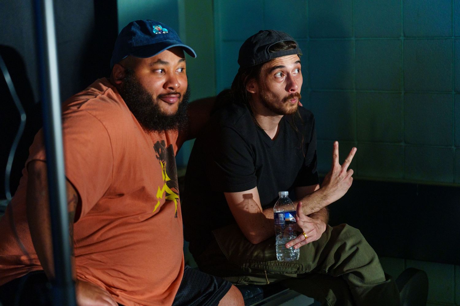 Two men sitting together in a dimly lit room, one holding a water bottle, both making casual gestures.