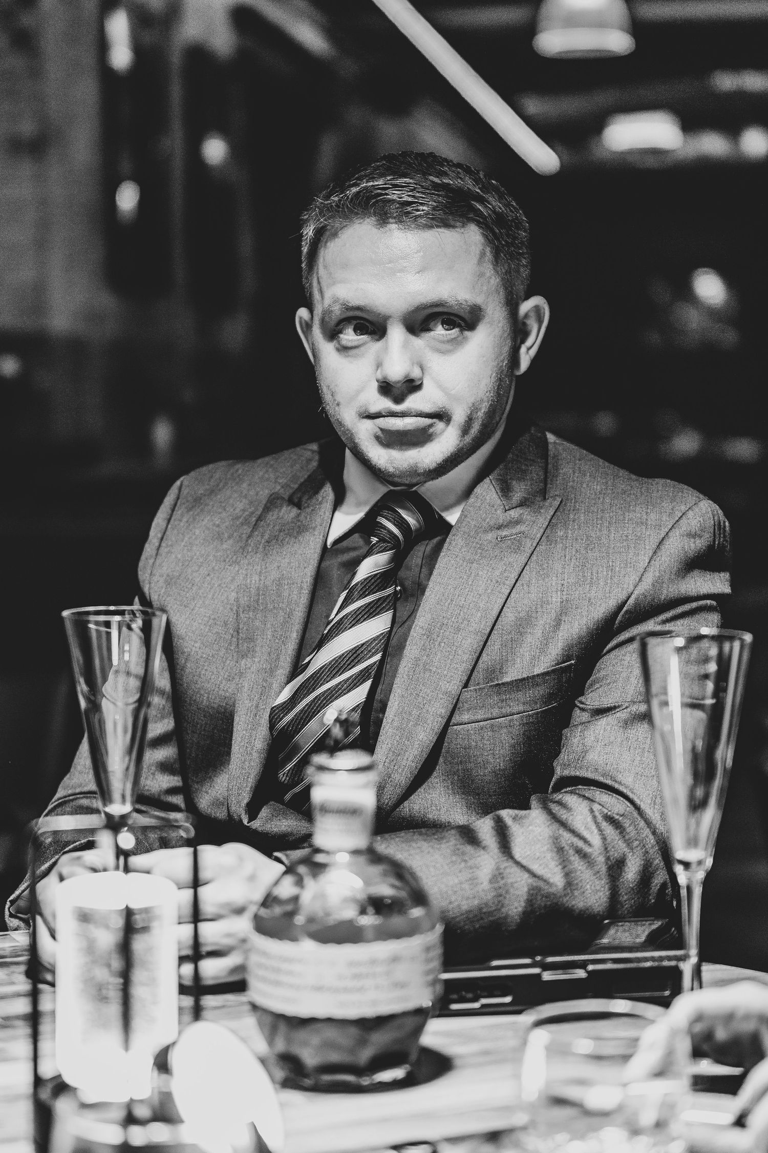 Black and white portrait of a person in business attire seated at a restaurant table with wine glasses.