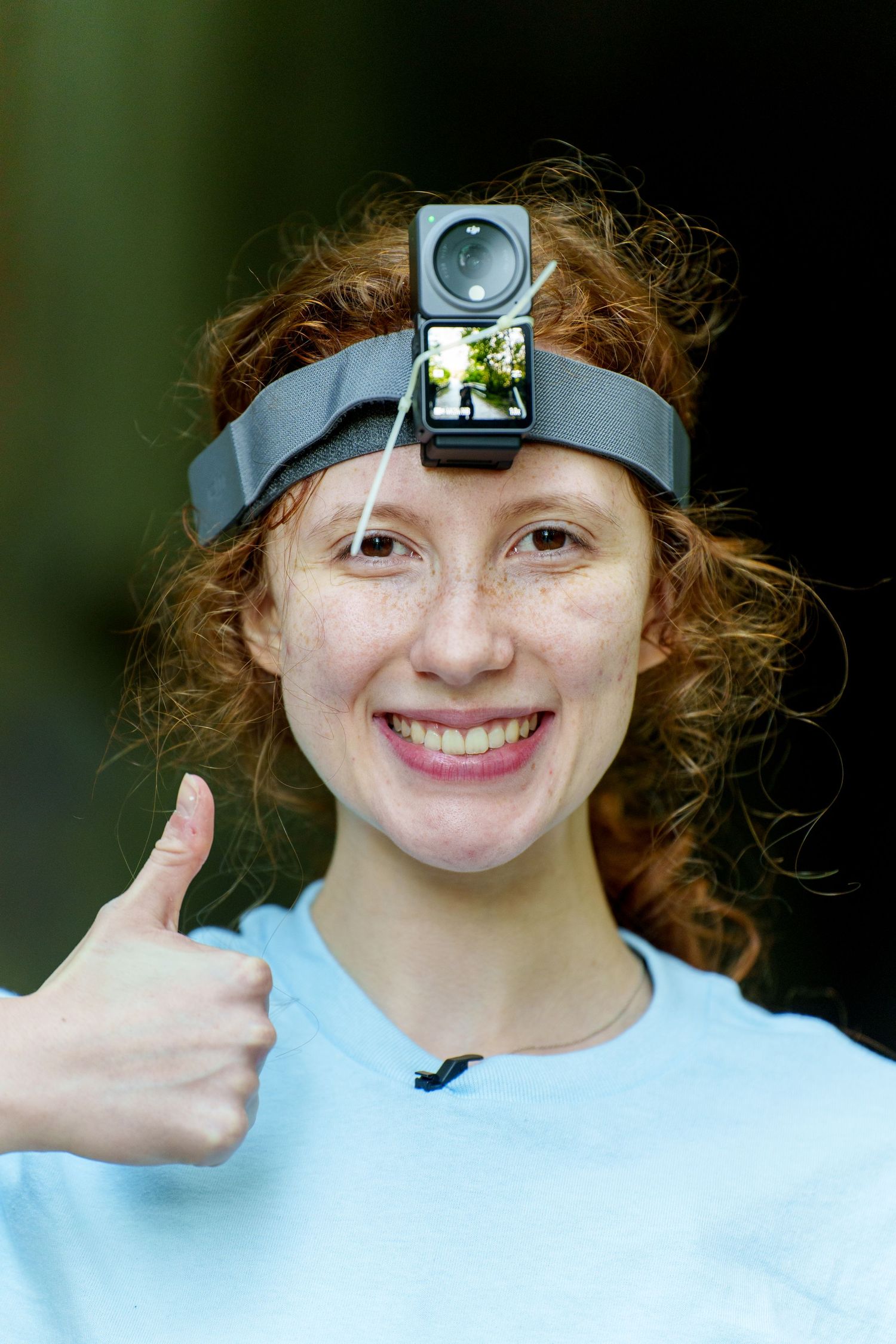Smiling redhead woman wearing a head-mounted action camera giving a thumbs up outdoors.