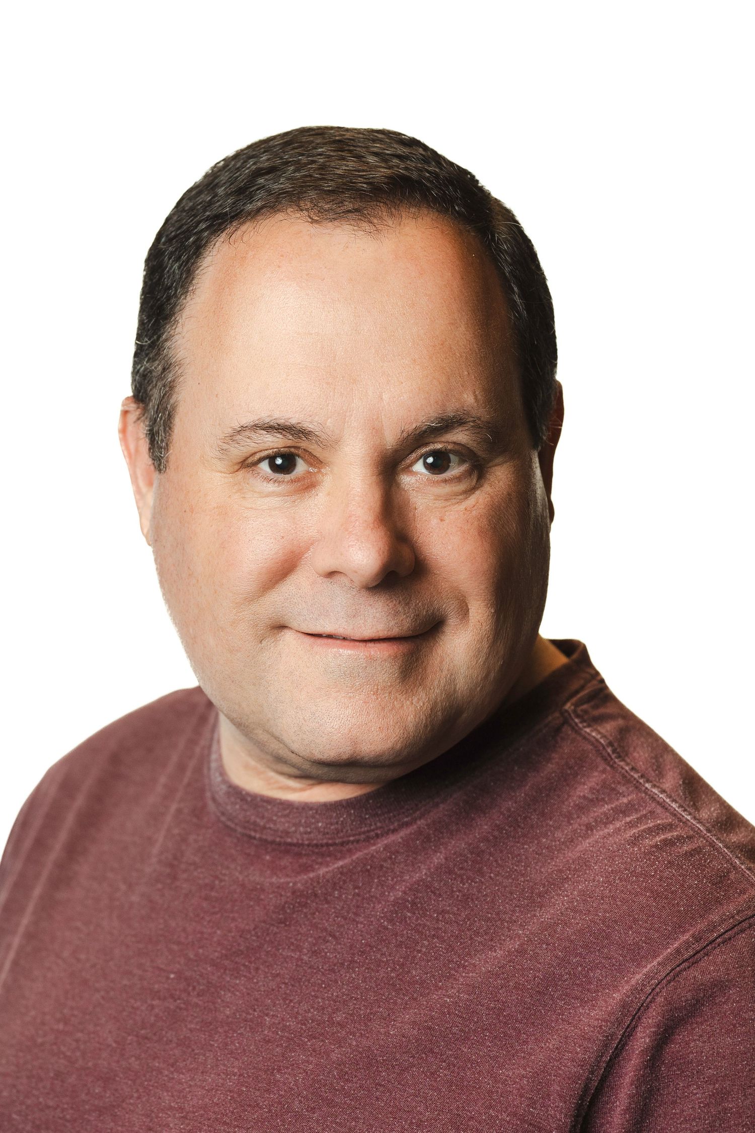 Middle-aged man with dark hair smiling softly, wearing a burgundy t-shirt against a white background.