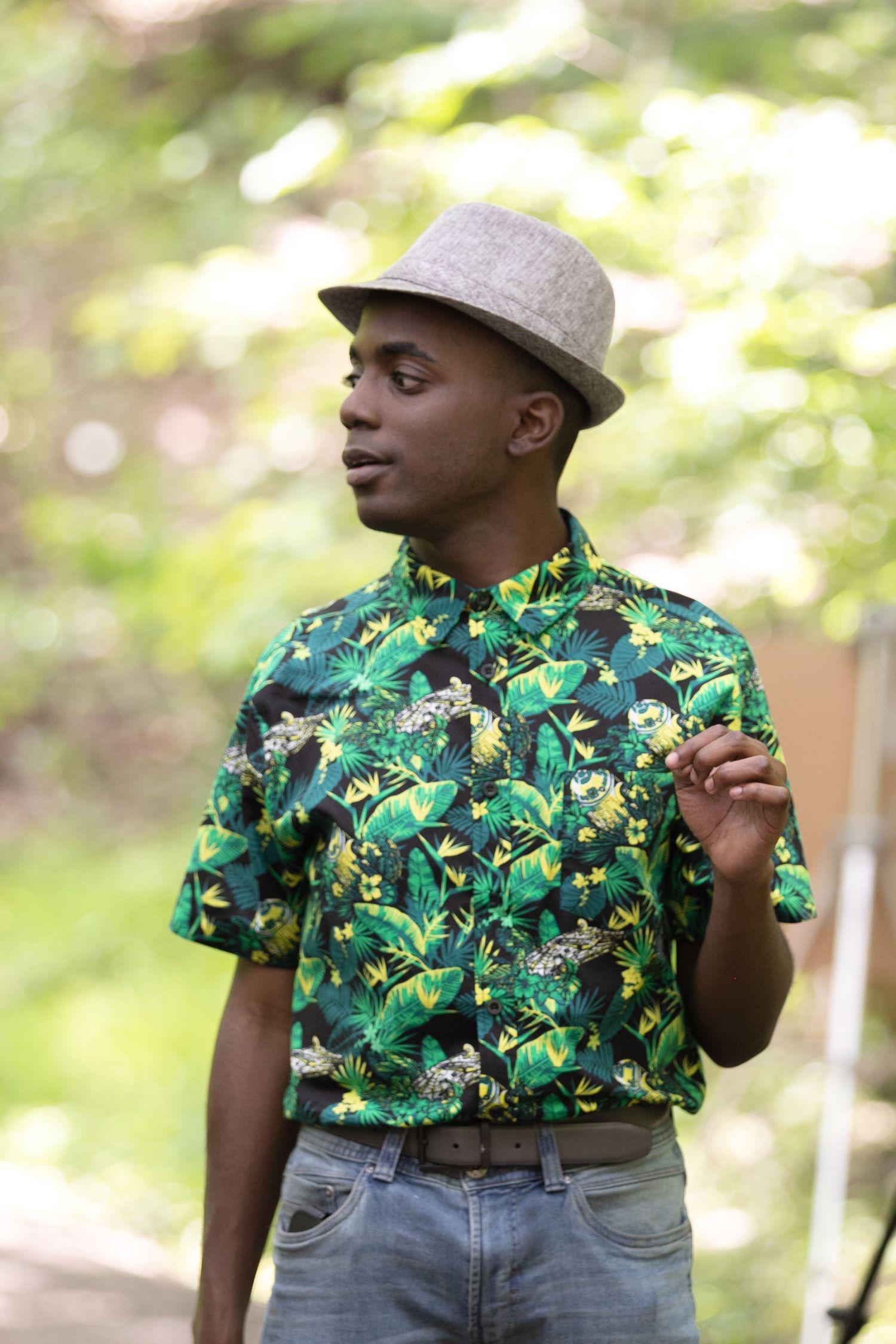 Young Black man in tropical floral shirt and gray fedora hat standing outdoors in green natural setting.