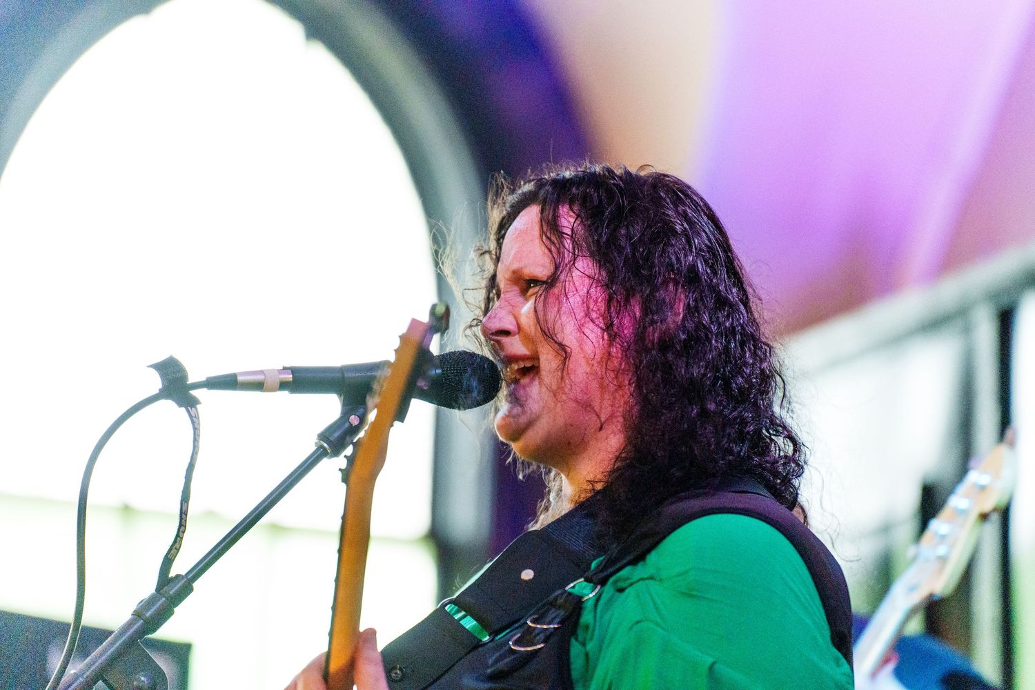 Long-haired guitarist in green shirt performs passionately at outdoor music festival stage.