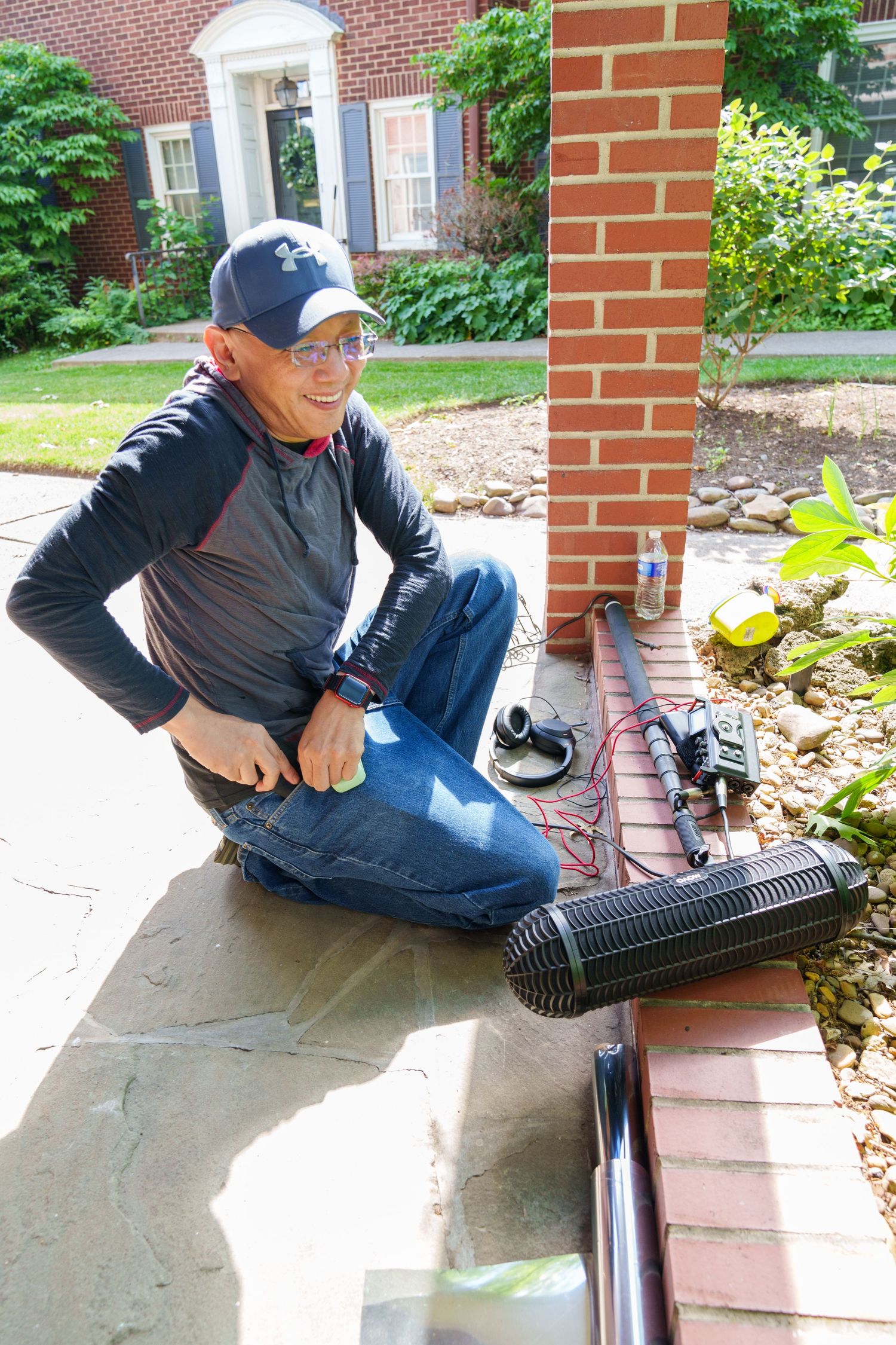 Man in cap crouching by brick pillar installing drainage system with black corrugated pipe on patio.