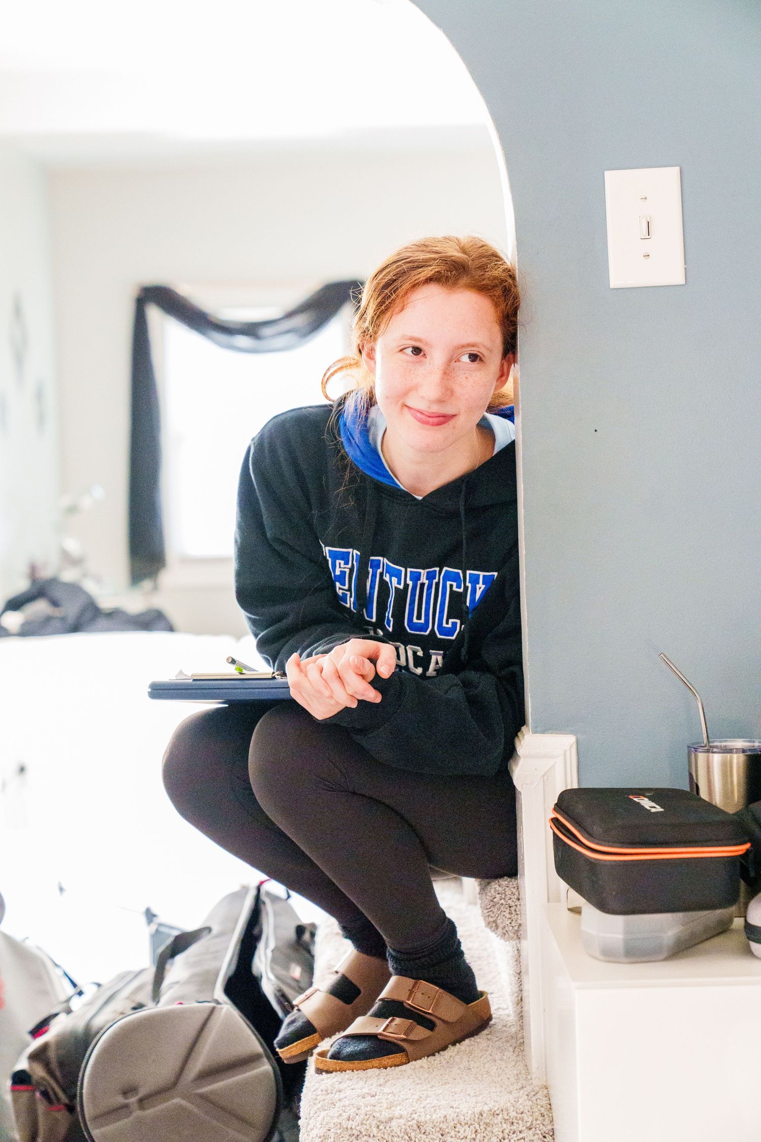 Young woman in Kentucky hoodie crouching by an archway holding a tablet indoors.