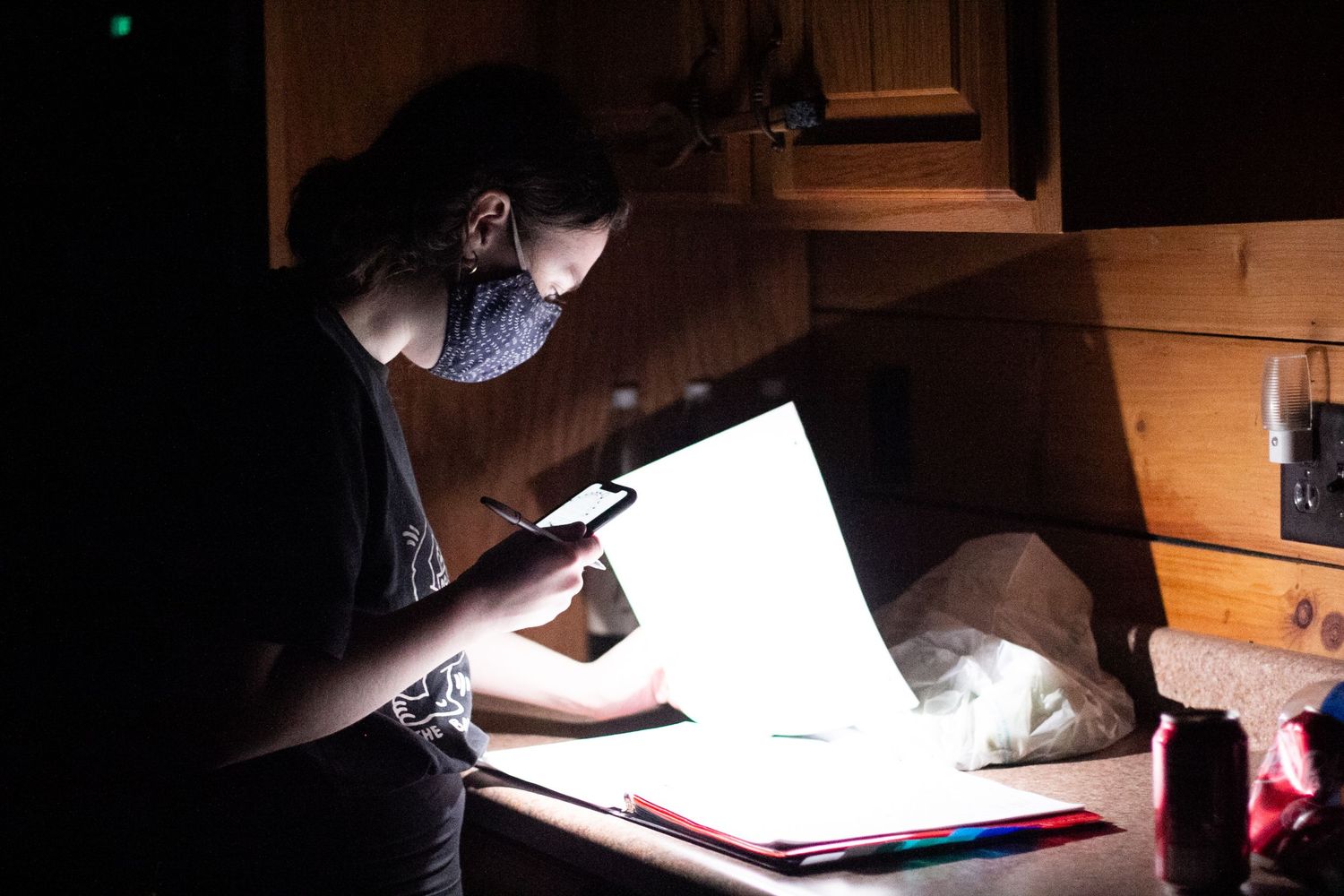 Person wearing a mask examines documents on a light box in a dimly lit wooden cabin at night.
