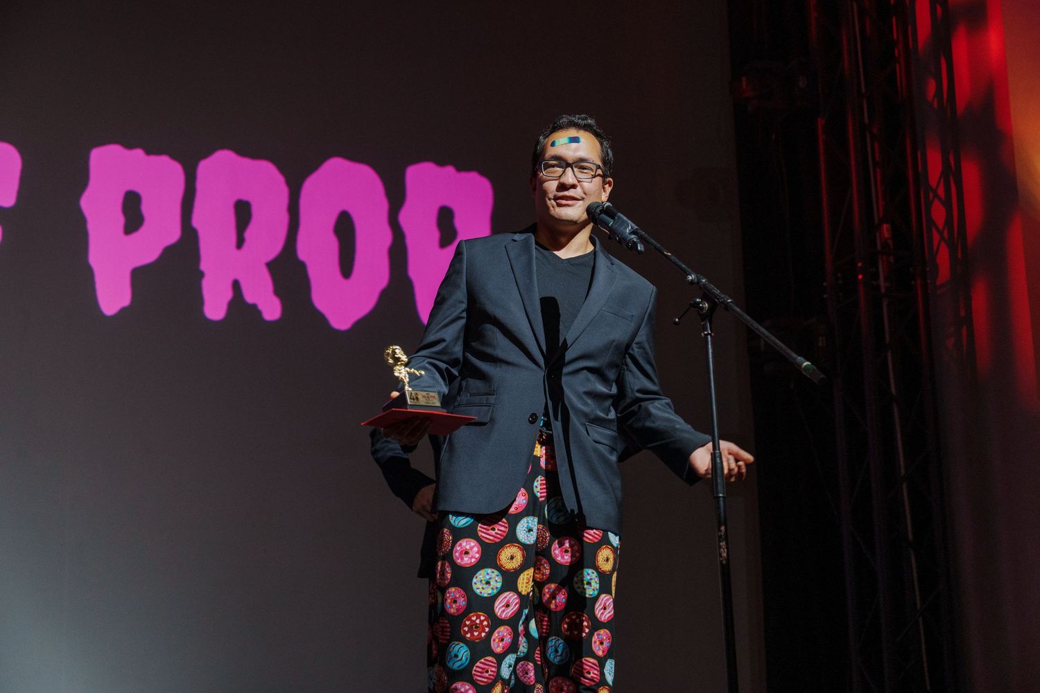 Speaker presenting on stage with pink text projection and dark curtain backdrop.