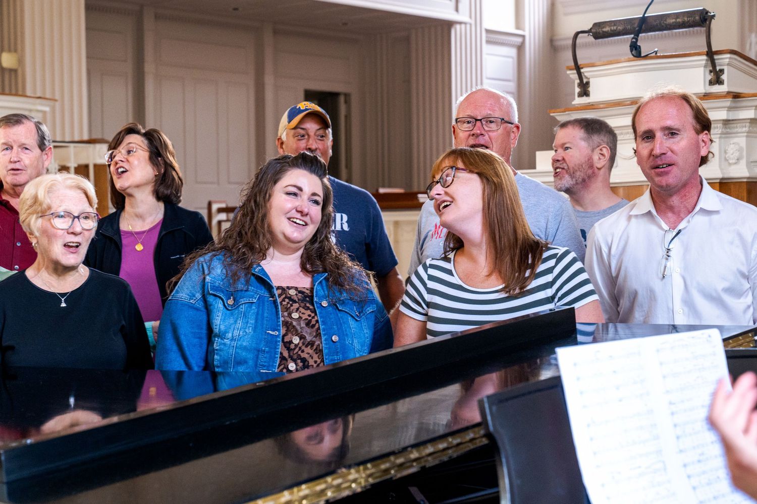 A joyful choir group singing together around a piano in a bright church hall setting.