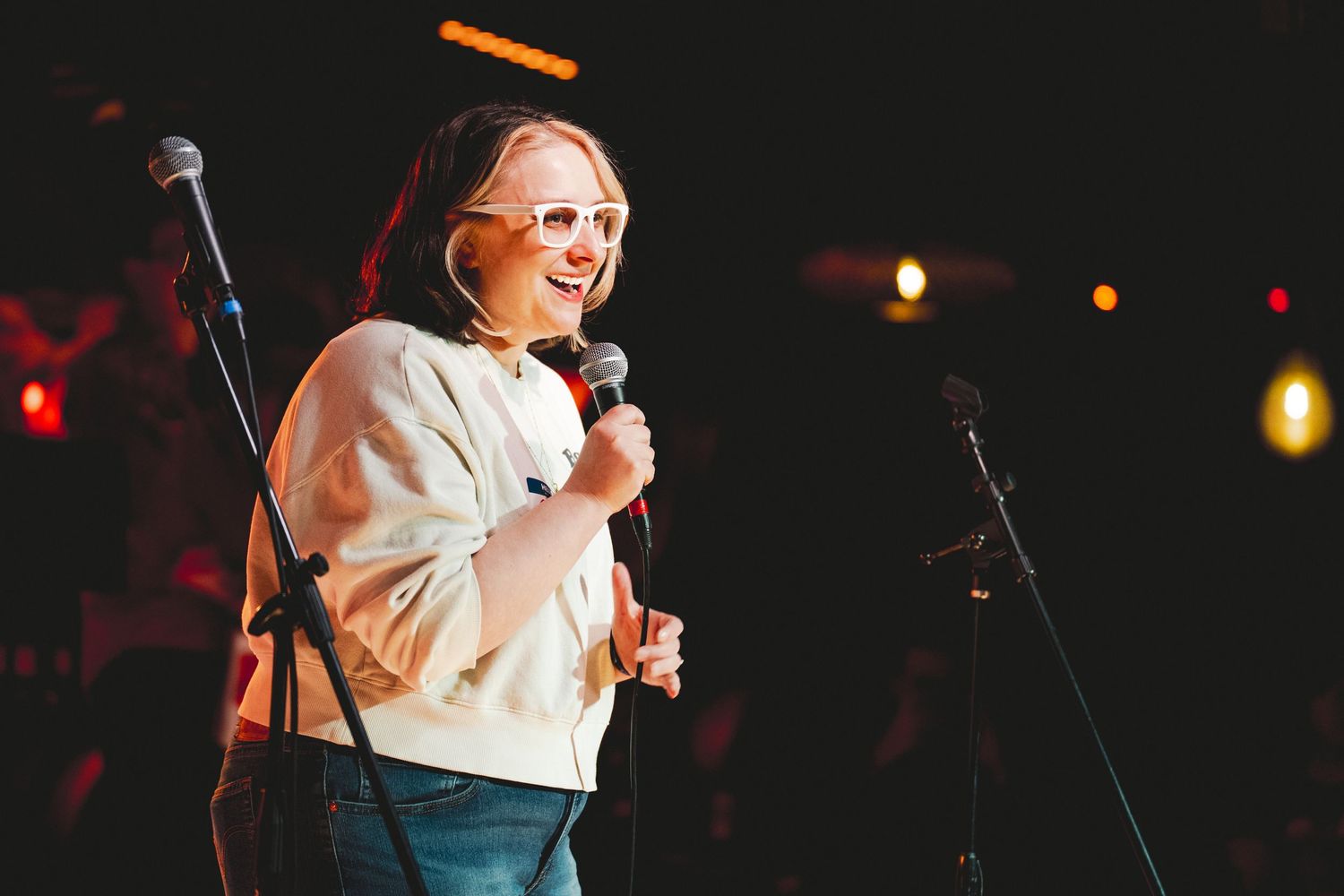 Smiling comedian performs stand-up on stage holding microphone, wearing white sweater and glasses under warm stage lighting.