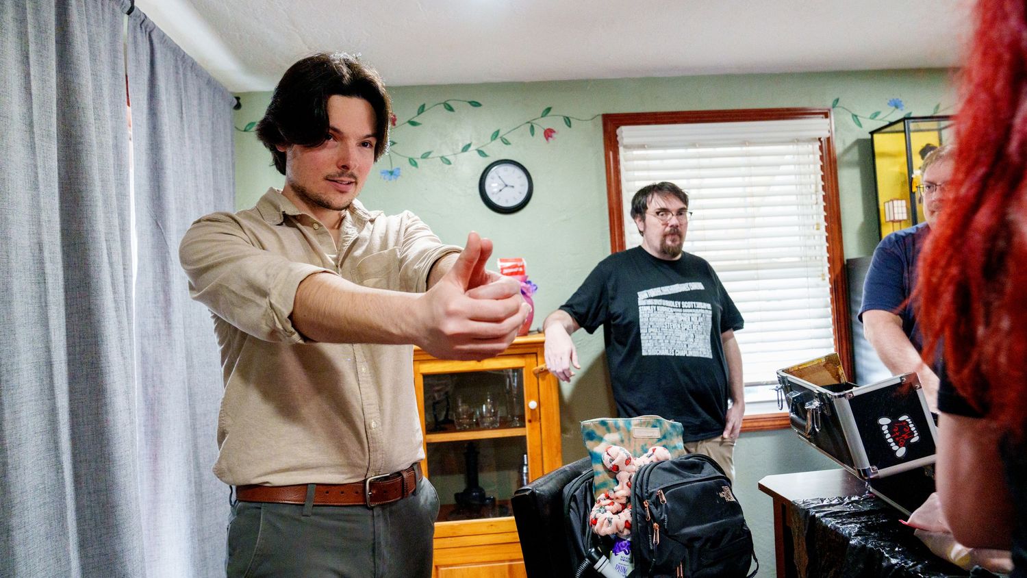 Man demonstrating magic trick indoors while two people watch in a decorated room with curtains and clock.