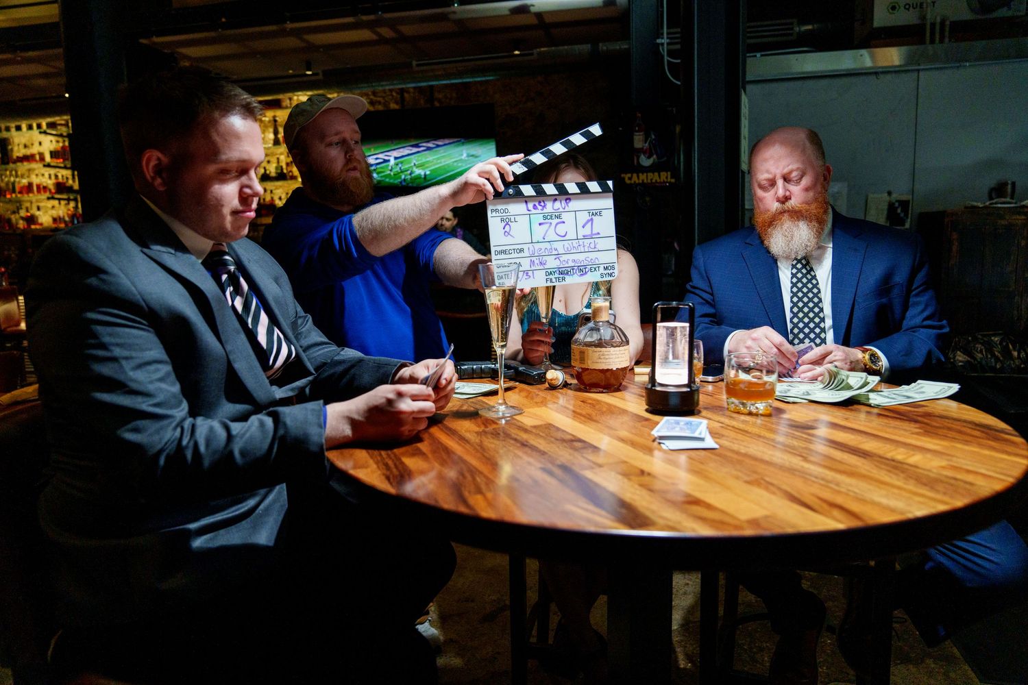 Two men in suits sit at a table during a film shoot, with a clapperboard held above them in a restaurant.