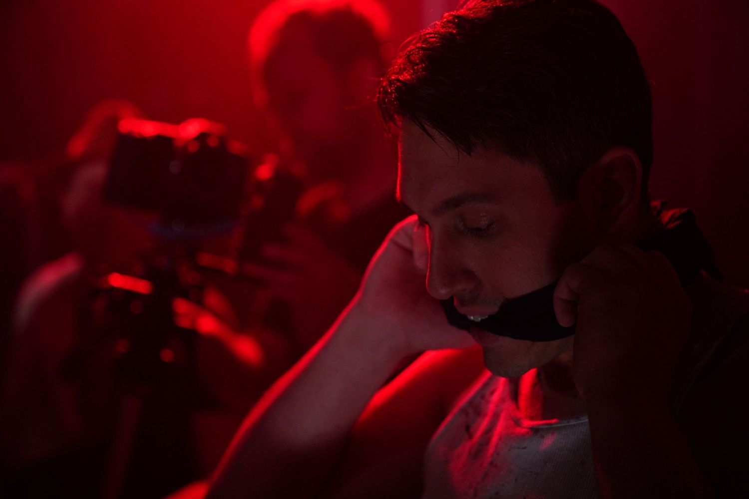 A young man in red-lit room holds phone to ear with camera operator visible in background.
