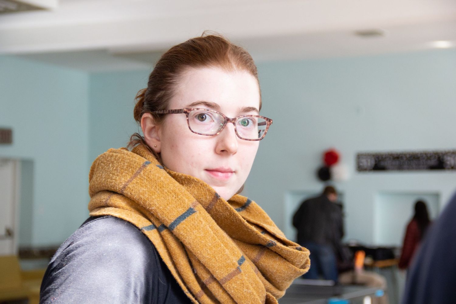 Young woman with glasses and mustard yellow scarf looks over shoulder indoors.