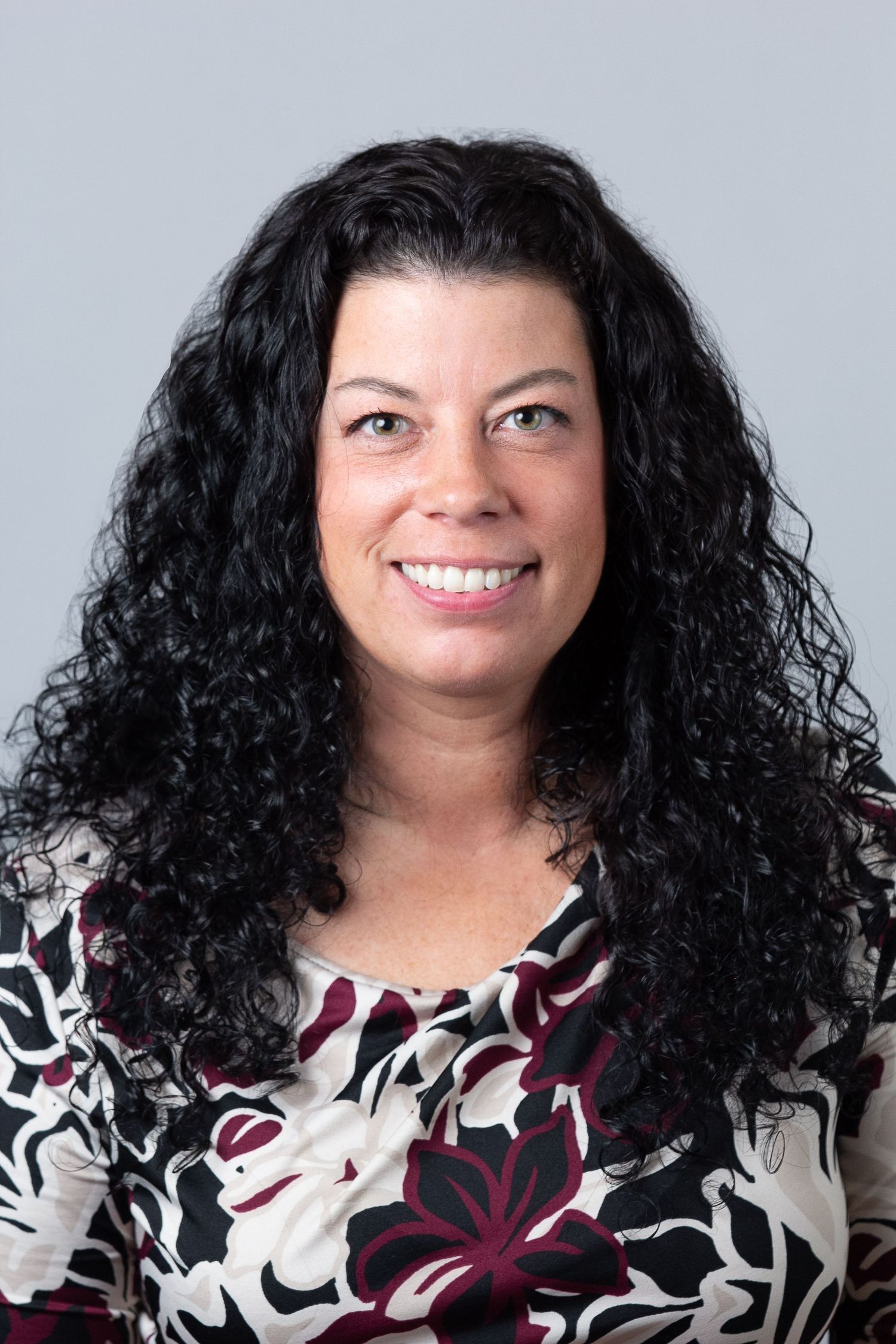 Professional headshot of a person with long dark curly hair wearing a floral print blouse against a light background.