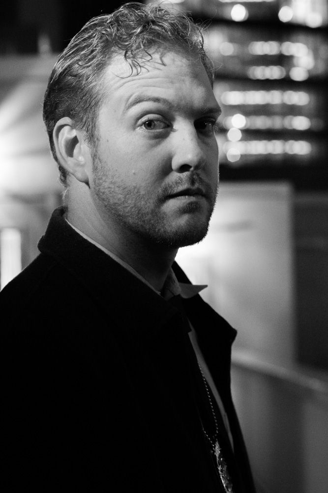 Black and white portrait of a man in a dark jacket with a necklace, looking over his shoulder at night.