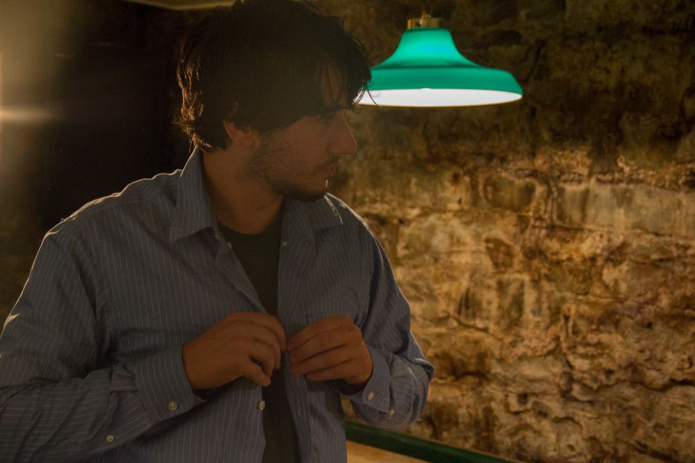 Man in blue striped shirt adjusting buttons under green pendant lamp in dimly lit cave setting.