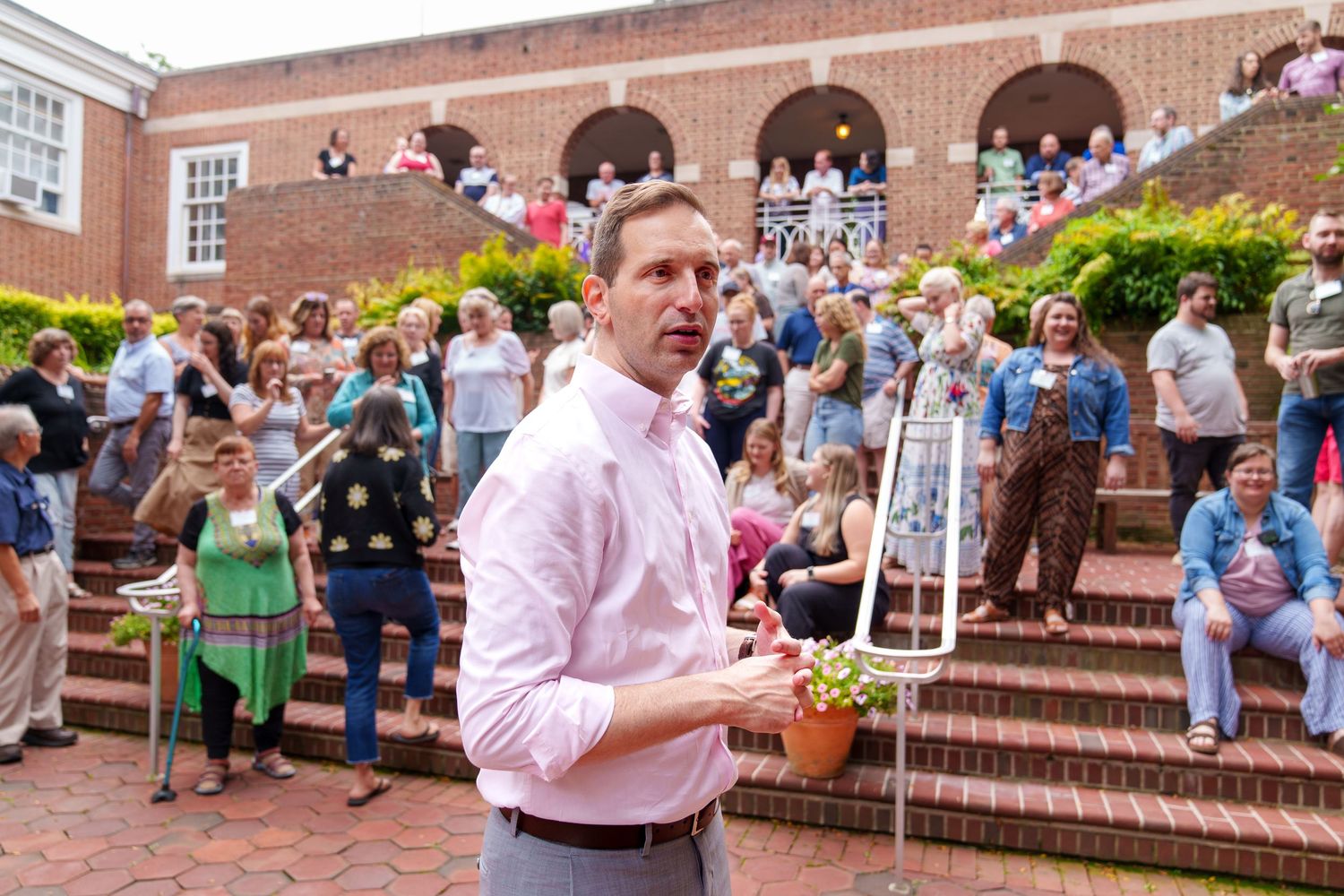 Man in pink shirt holds potted plant outdoors, addressing a large crowd gathered on brick steps.